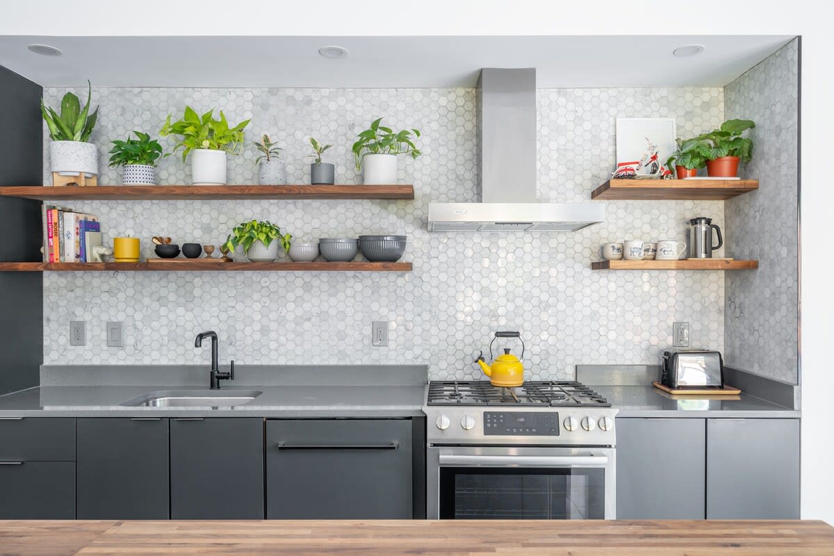 A detailed view of the kitchen shows grey countertops, a stainless steel gas range with a hood, a sink with a black faucet, and open wooden shelves adorned with potted plants and dishware against a white hexagonal tile backsplash.