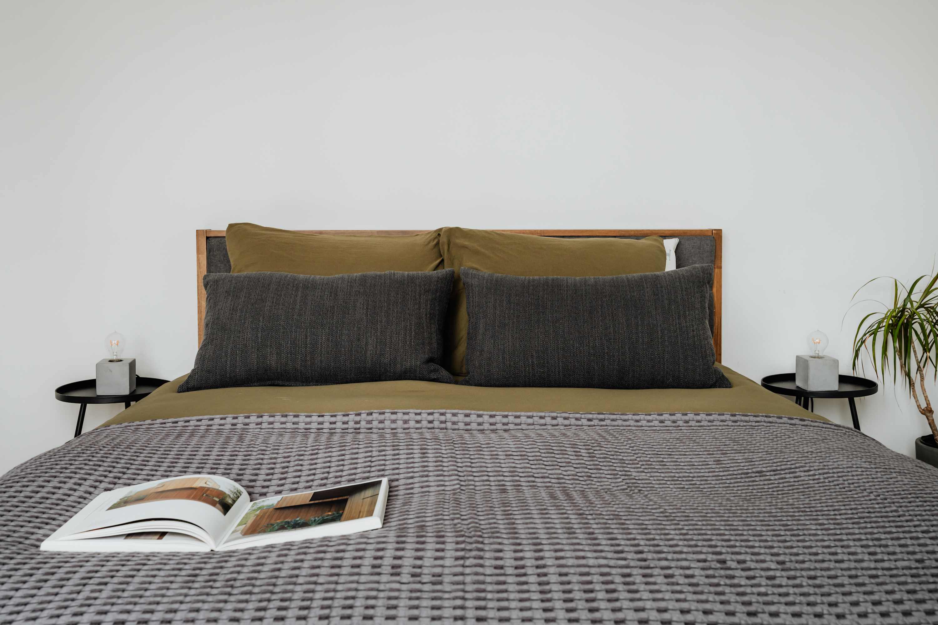 A close-up view of a bed with a wooden headboard, dressed in olive green sheets and dark gray rectangular pillows, topped with a gray textured blanket and an open magazine. Small concrete side tables with minimalist lamps flank the bed, set against a clean white wall.