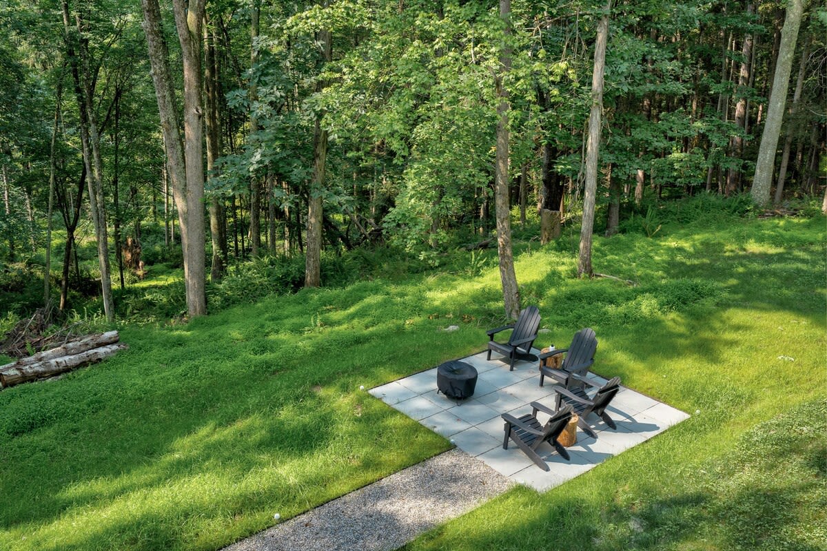 An aerial view showcases an outdoor fire pit area on light gray pavers, surrounded by a vibrant green lawn and dense forest. Four black Adirondack chairs are arranged around a black fire pit, with a gravel path leading to the paved seating area.