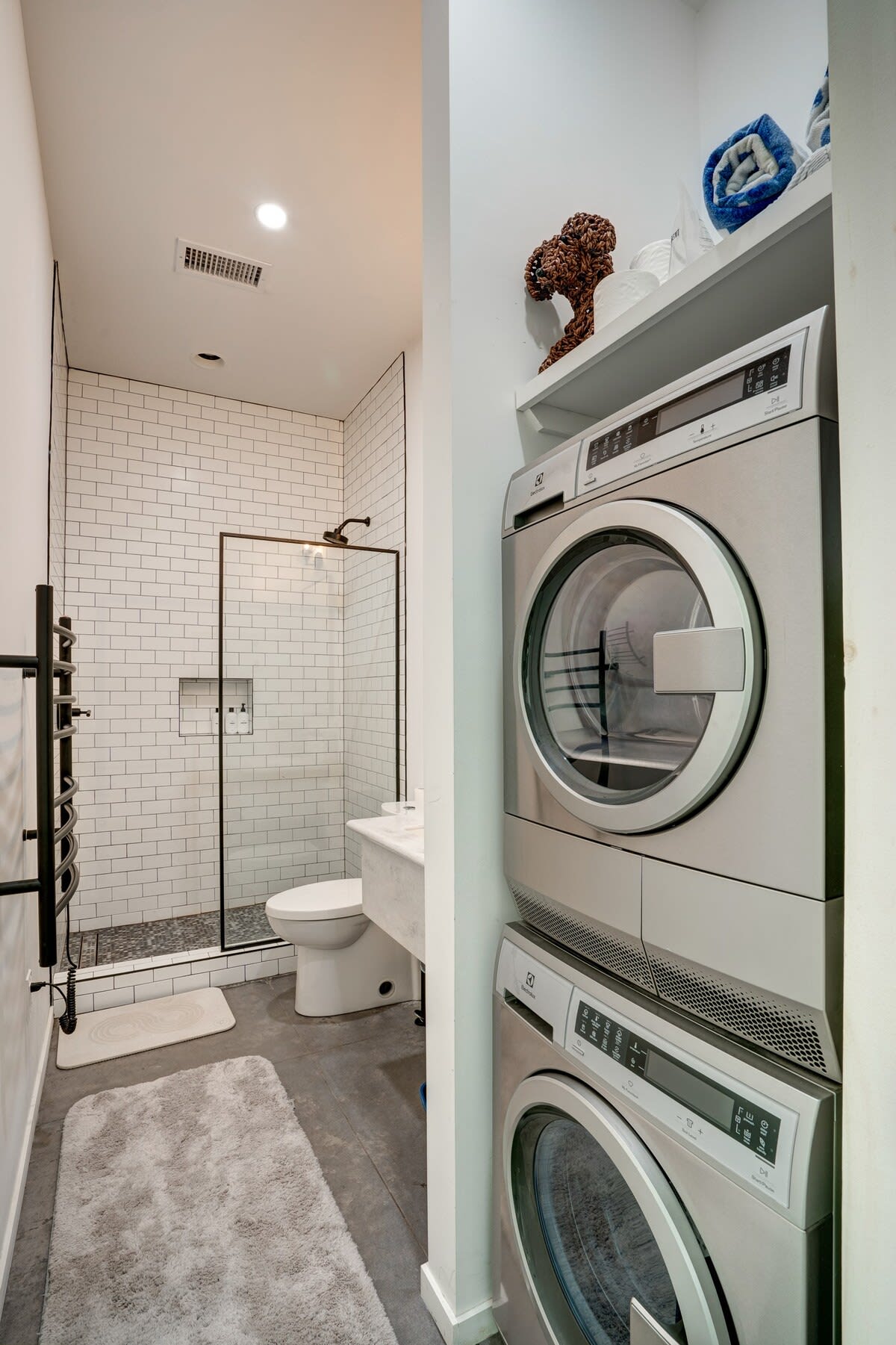 A narrow, modern bathroom features a white subway tile shower with a glass door and black frame, a toilet, and a vanity. Adjacent to this, a stacked washer and dryer unit is built into an alcove with white shelving above, creating a combined bathroom and laundry space.