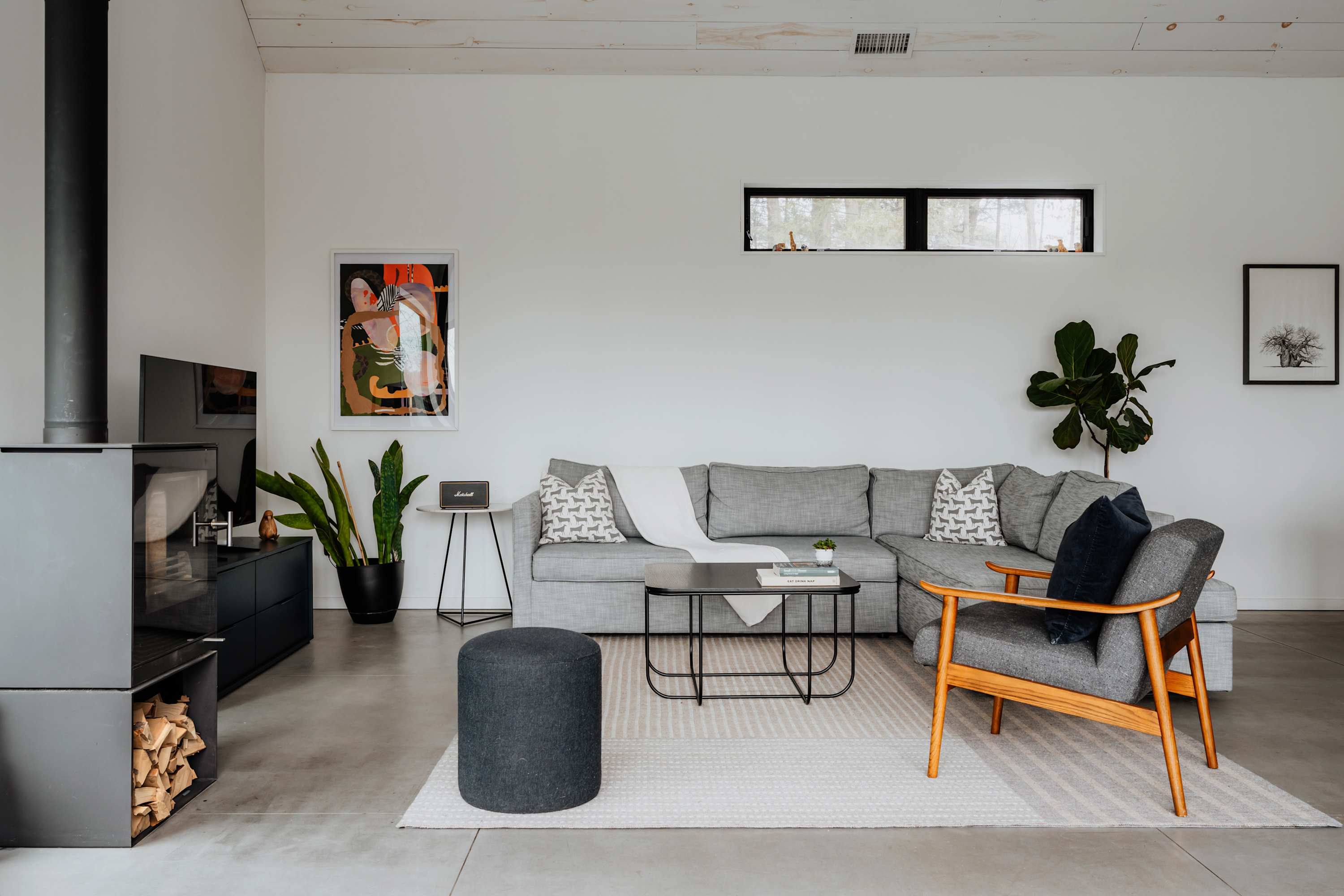 A contemporary living room showcases a large grey sectional sofa, a wooden armchair, a black metal coffee table, and a modern wood-burning stove, all set on a light patterned rug over concrete floors.