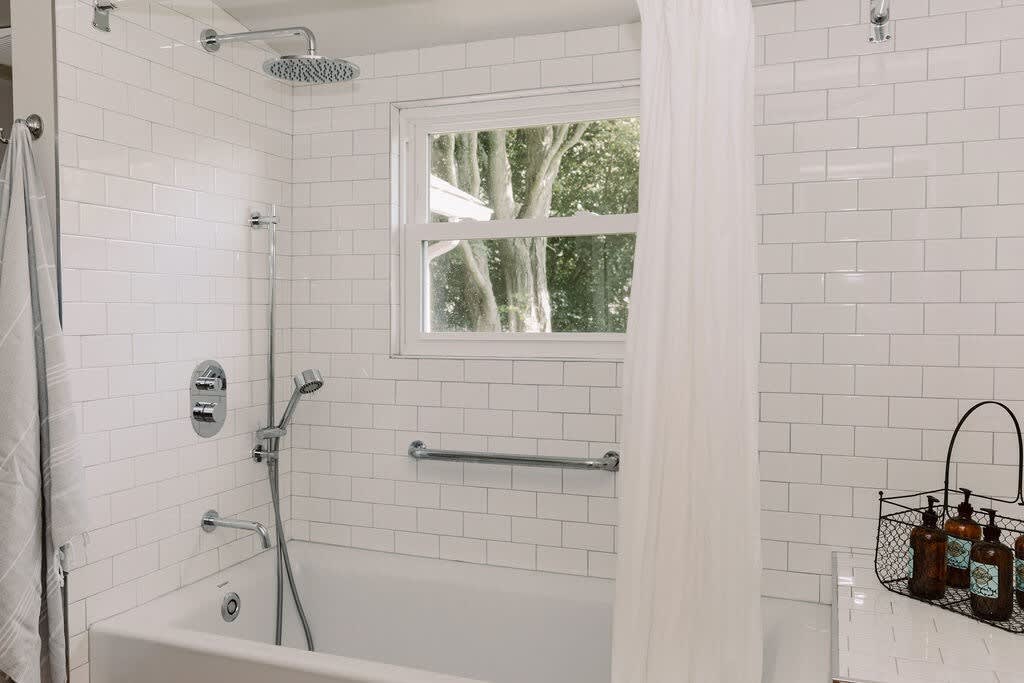 A white subway-tiled shower and bathtub area with a rain shower head and a handheld shower, featuring a window with a view of trees. A white shower curtain and grab bar are also visible.