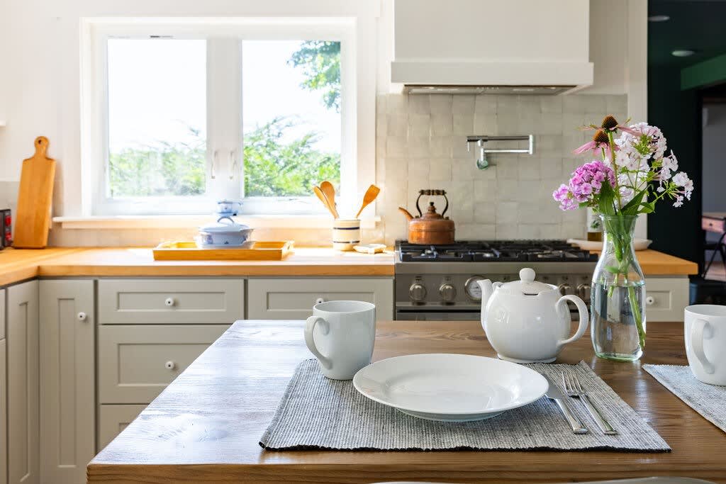 Kitchen counter with light wooden butcher block countertops, a white tiled backsplash, a gas range with a pot filler, and a window overlooking greenery, set with a teapot and floral arrangement.