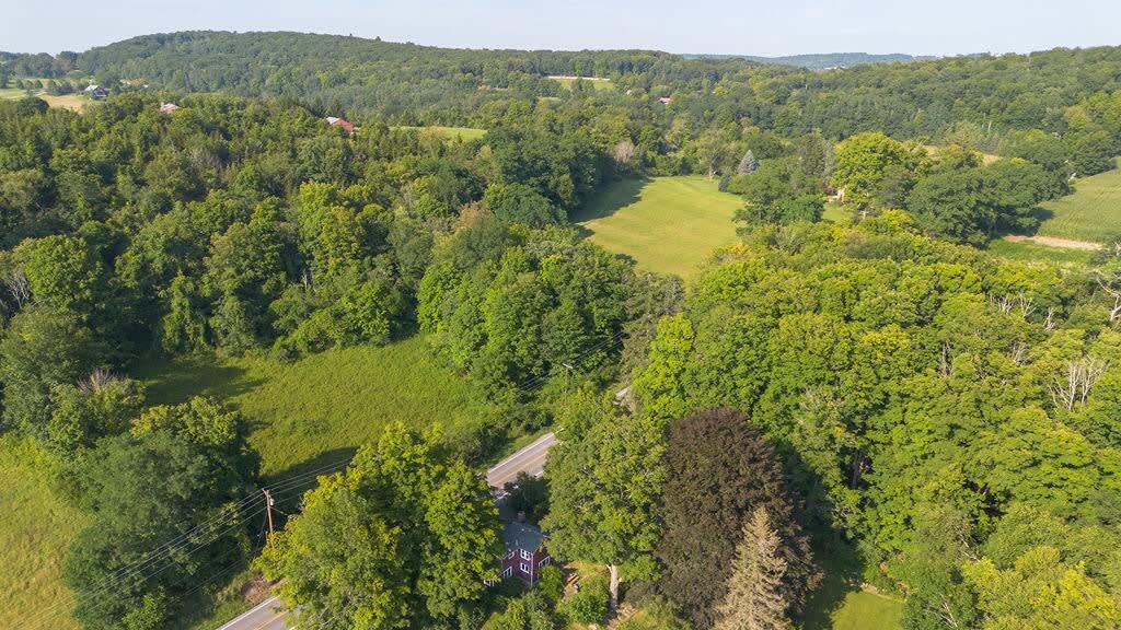 An aerial view reveals the red farmhouse surrounded by vast expanses of lush green forests and fields, with a winding road nearby, highlighting its secluded and natural rural location.