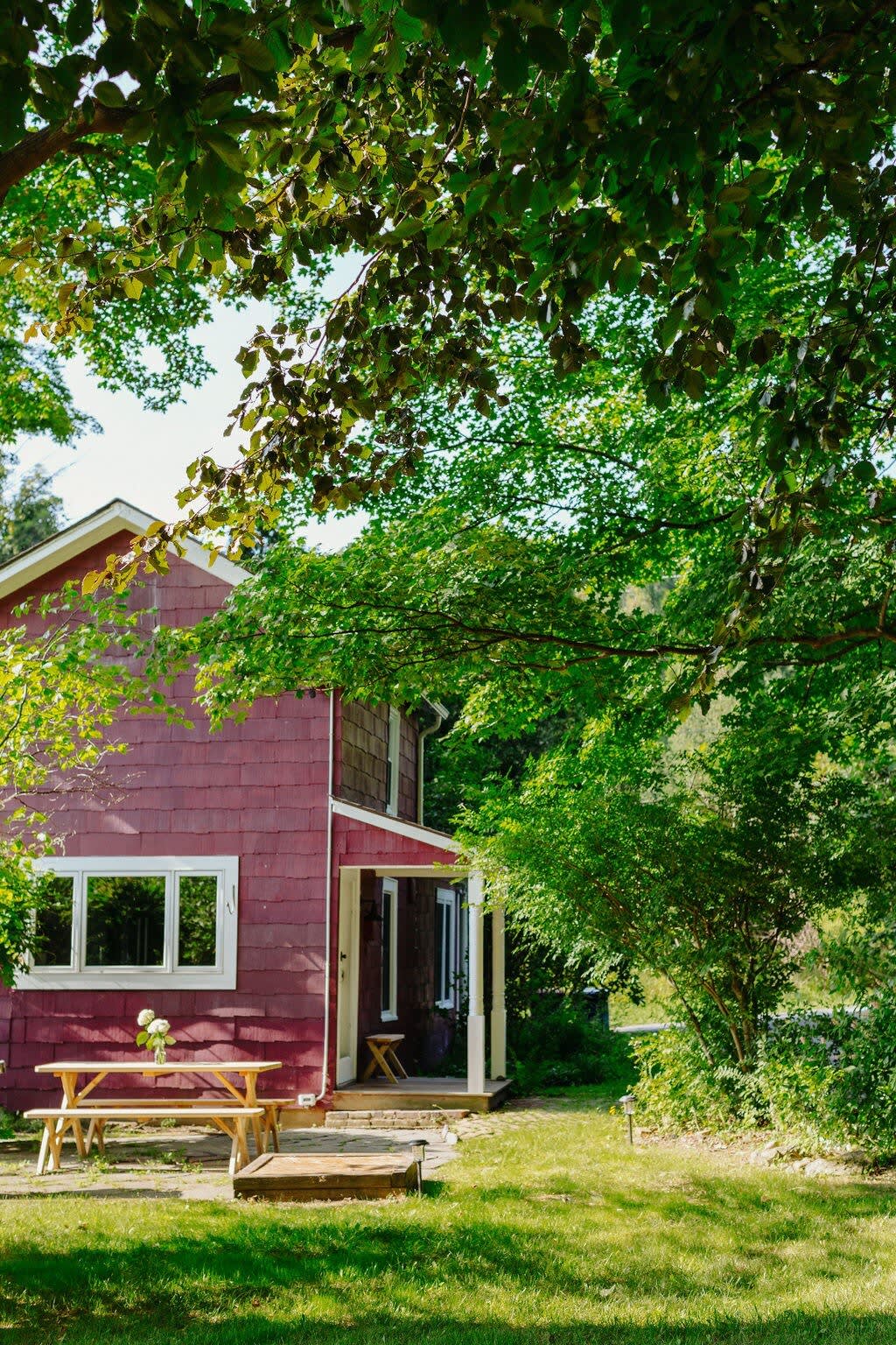 The red farmhouse is nestled among vibrant green trees and a grassy lawn, featuring a wooden picnic table on a stone patio and a covered porch entrance, showcasing a serene rural setting.