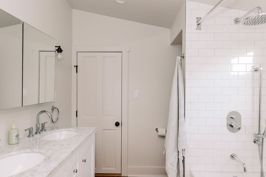 A modern bathroom with a double vanity featuring white sinks and a marble countertop, large mirrors, and a white subway-tiled shower/tub combo. A white paneled door is visible in the background.