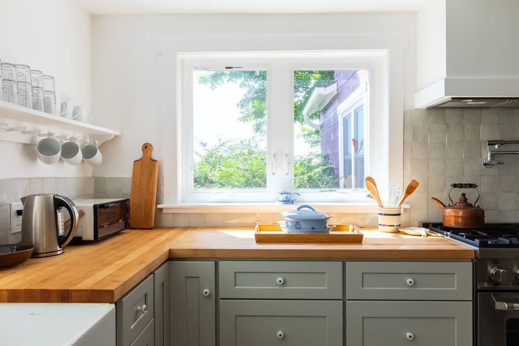 Bright kitchen counter with light wooden butcher block countertops, a white farmhouse sink, a kettle, toaster oven, and open shelving with glassware, illuminated by a large window.