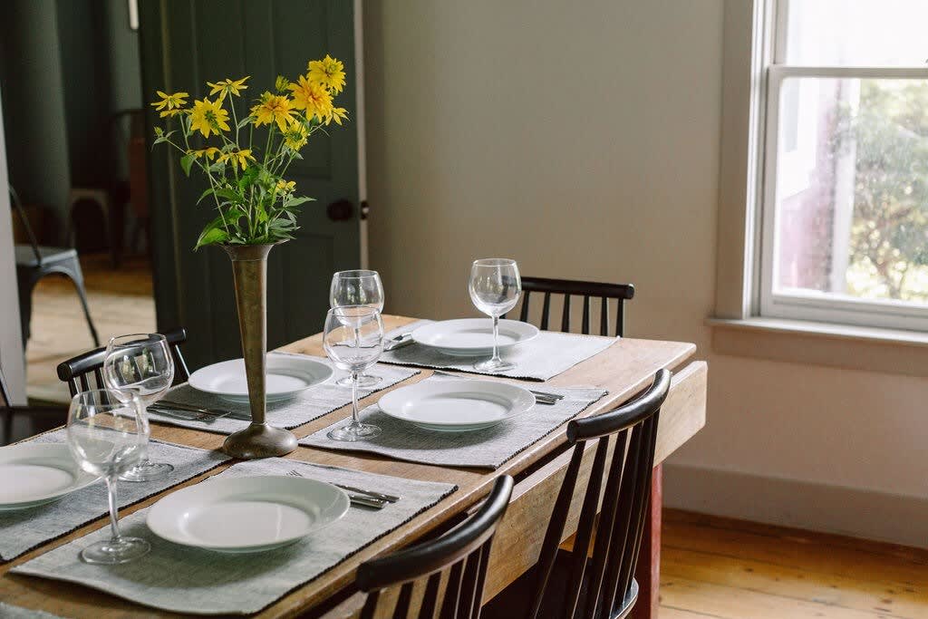 Dining area with a rustic wooden table set for four with white plates and wine glasses, surrounded by dark wooden chairs, next to a window, with a vase of yellow flowers as a centerpiece.