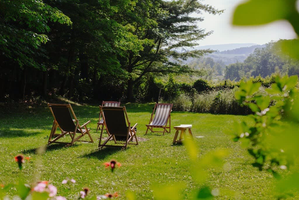 Lush green lawn with several wooden lounge chairs and a small side table, set against a backdrop of dense trees and distant rolling hills. The scene suggests a peaceful outdoor relaxation area.
