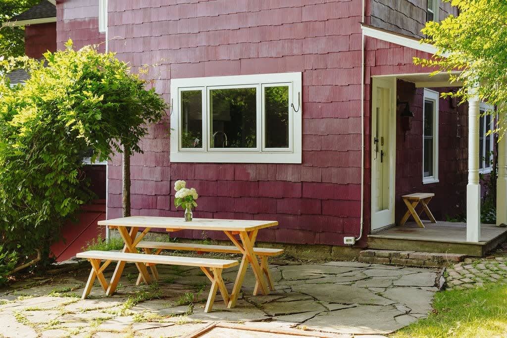 Outdoor patio area with a light wood picnic table and benches on a stone slab surface, adjacent to the red-sided house. Lush green foliage surrounds the space, offering a pleasant spot for al fresco dining.