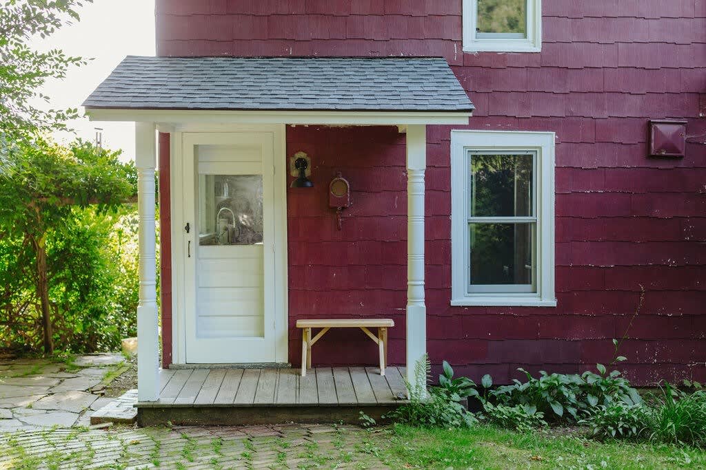 The front entrance of the red farmhouse features a white screen door under a small covered porch with white pillars, a simple wooden bench, and a stone path leading up to it, surrounded by green foliage.