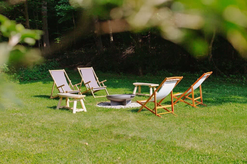 A wide shot of a spacious grassy yard with a central fire pit area surrounded by several wooden lounge chairs and small side tables. The area is backed by a dense line of trees.