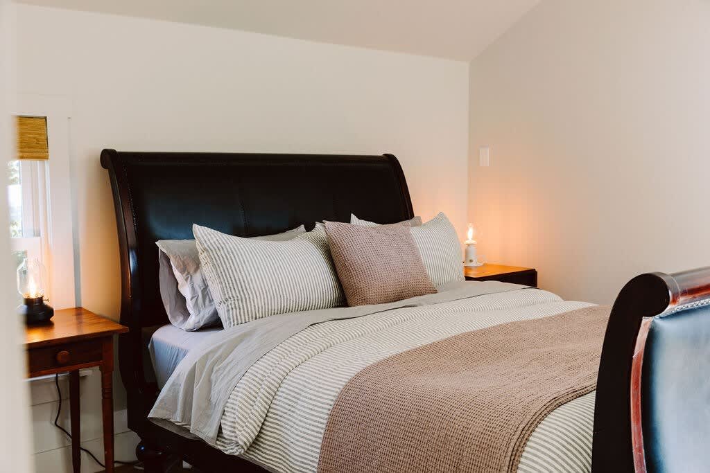A close-up of a dark brown leather sleigh bed with striped and pink bedding, flanked by wooden nightstands with lamps. A window with a woven blind is visible on the left, and a warm glow from a lamp illuminates the right side.