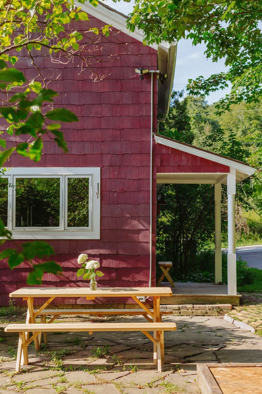 The side of the red farmhouse features a stone patio with a wooden picnic table and benches, adorned with a vase of white flowers, set against a backdrop of green trees and a glimpse of a road.