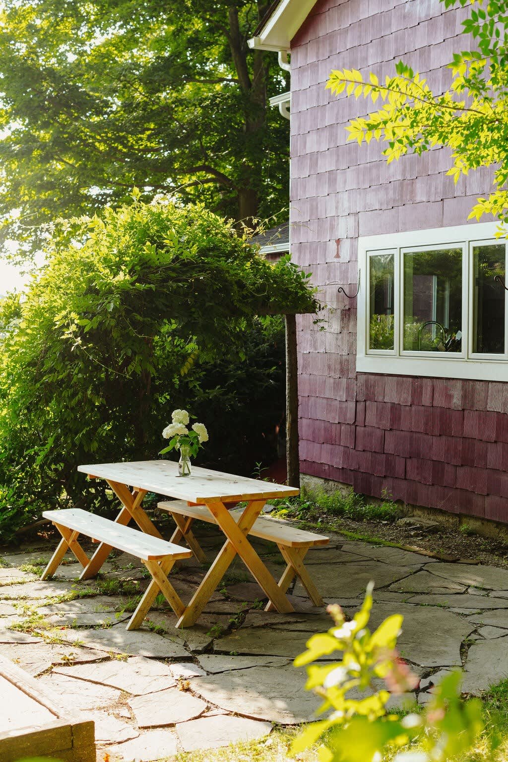 A bright stone patio next to the red farmhouse features a light wooden picnic table with matching benches and a vase of white flowers, surrounded by dense green bushes and trees under a sunny sky.