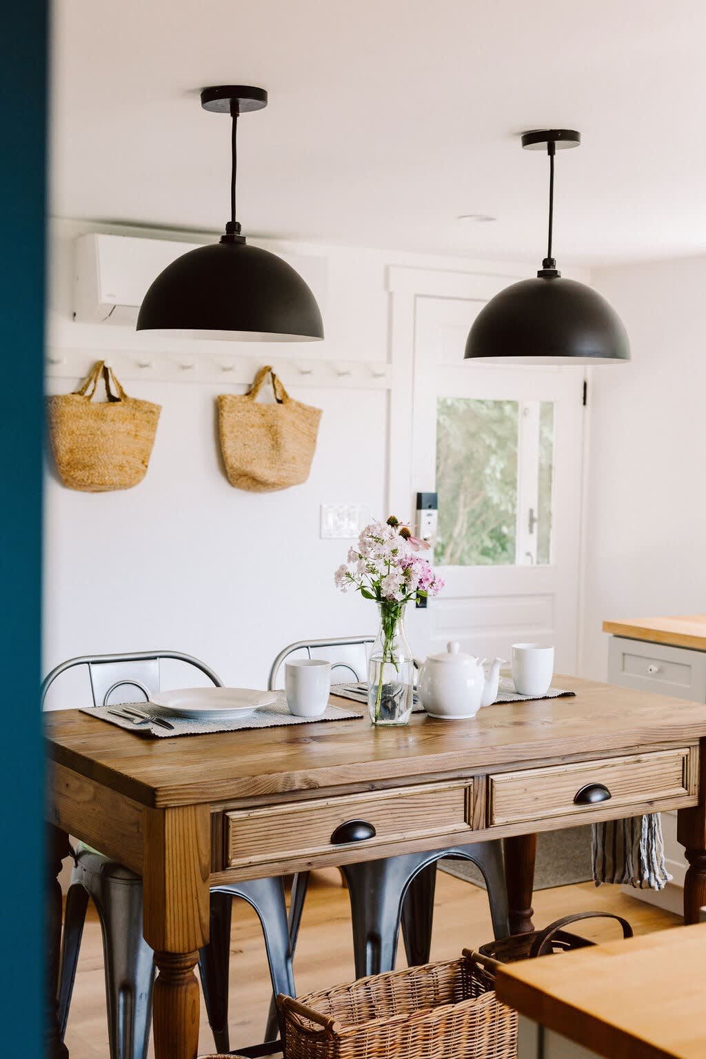 Rustic wooden dining table set with white dishes and a floral centerpiece, flanked by industrial metal chairs, beneath two black dome pendant lights, with straw bags hanging on a nearby wall.
