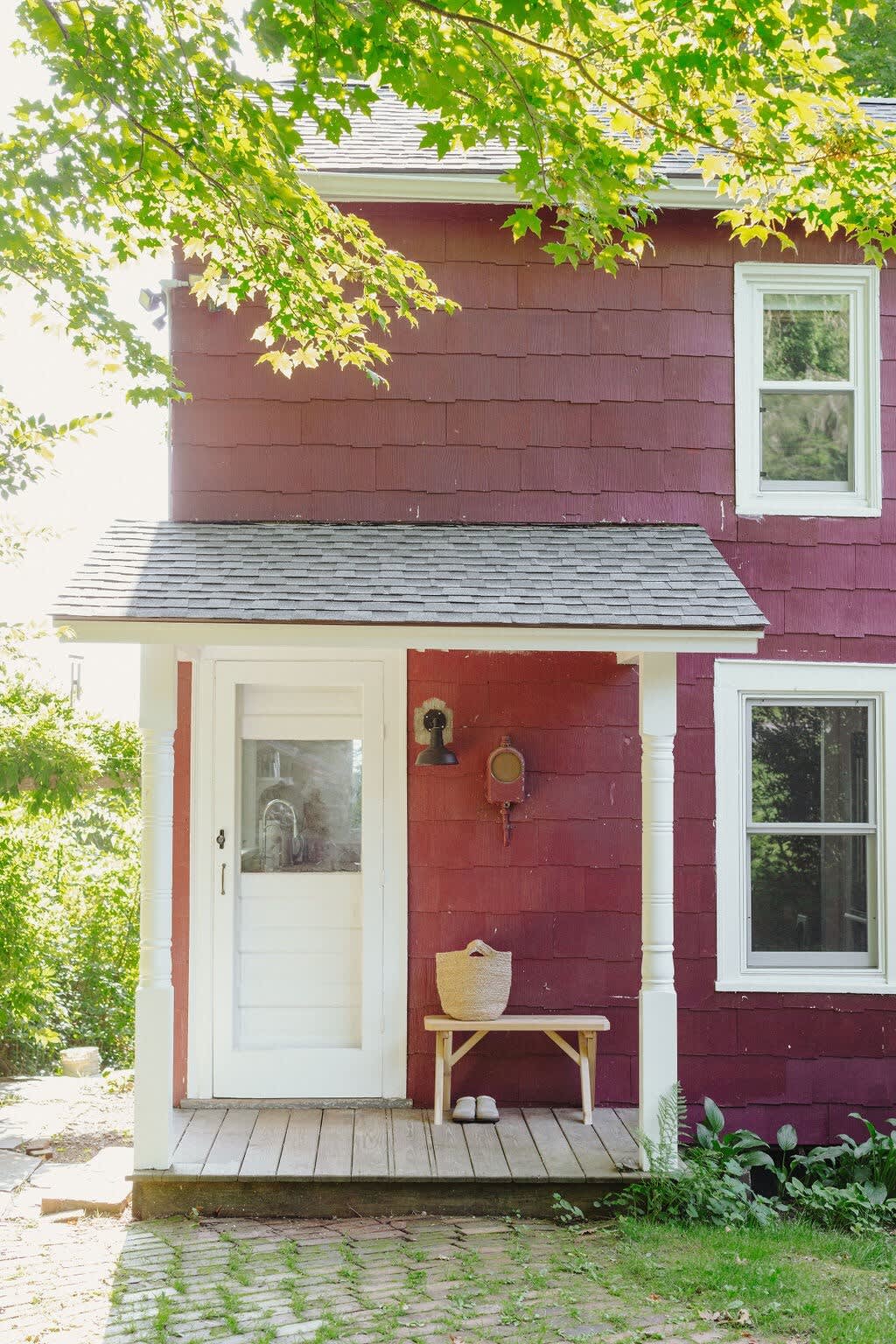 The front entrance of a charming red farmhouse features a white screen door under a small covered porch with white pillars, a wooden bench, and a woven basket, surrounded by lush greenery and a stone path.