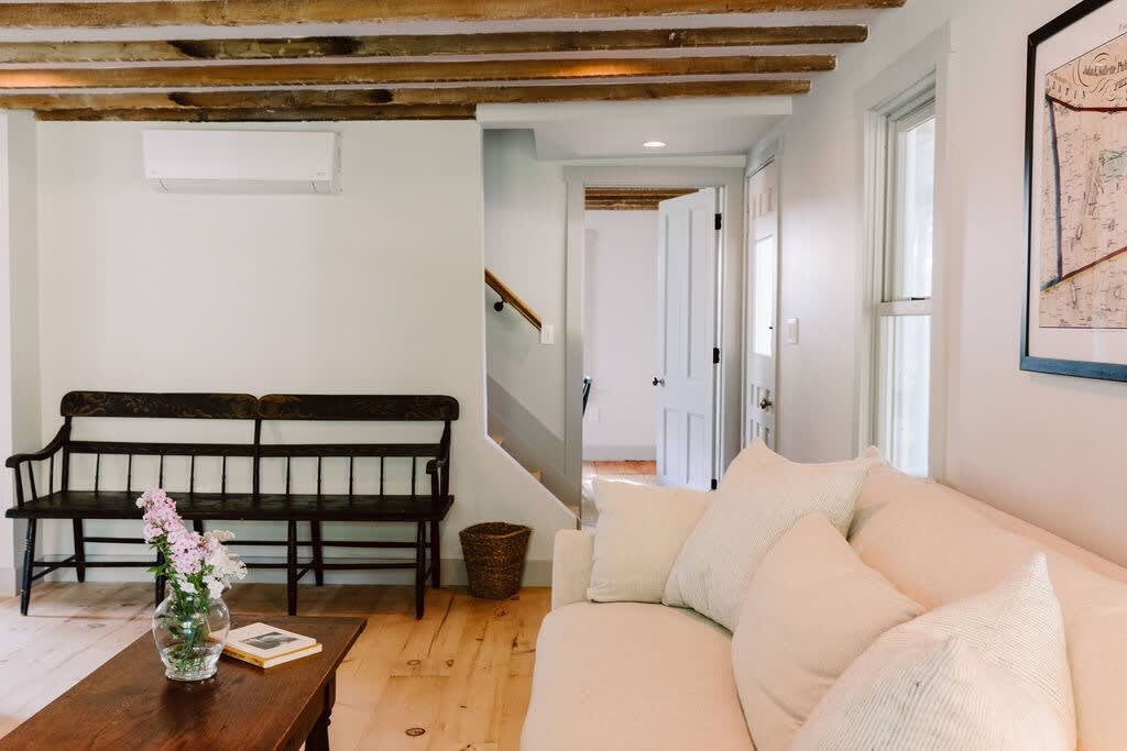 Cozy living room with a light-colored sofa, dark wooden coffee table, and a long dark wooden bench, featuring exposed wooden ceiling beams and light hardwood floors, with a mini-split AC unit on the wall.