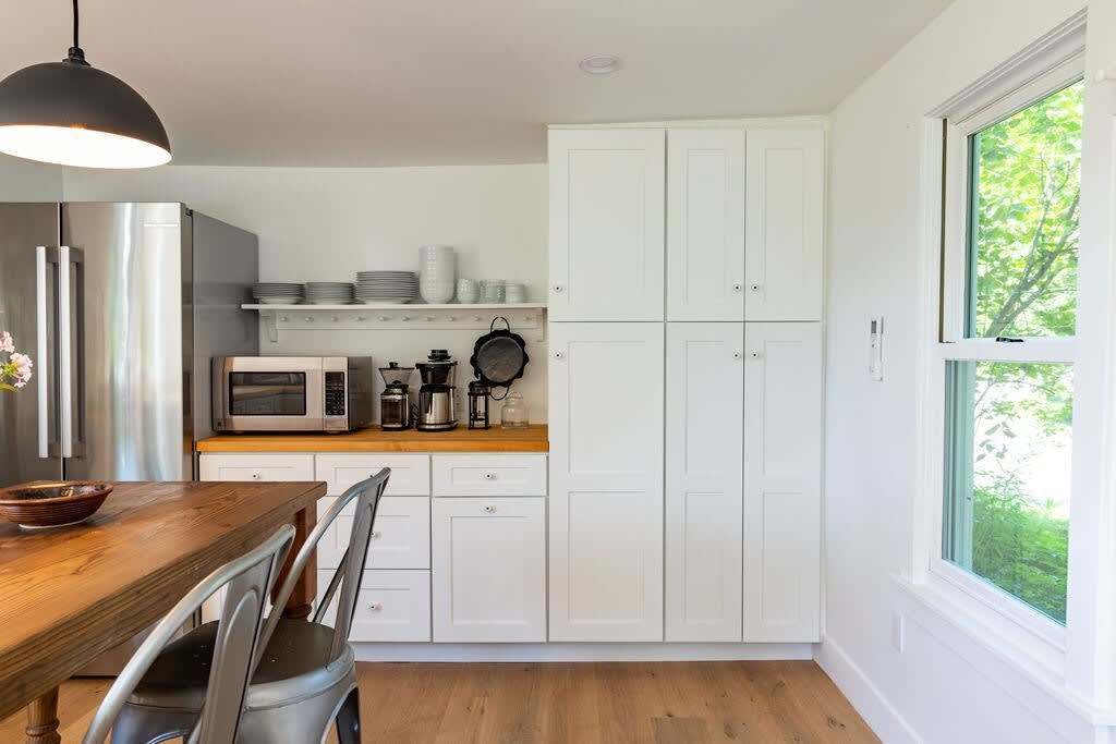 Bright kitchen area featuring a stainless steel refrigerator, microwave, and coffee maker on a wooden countertop, with white shaker-style cabinets and light hardwood flooring, next to a large window.