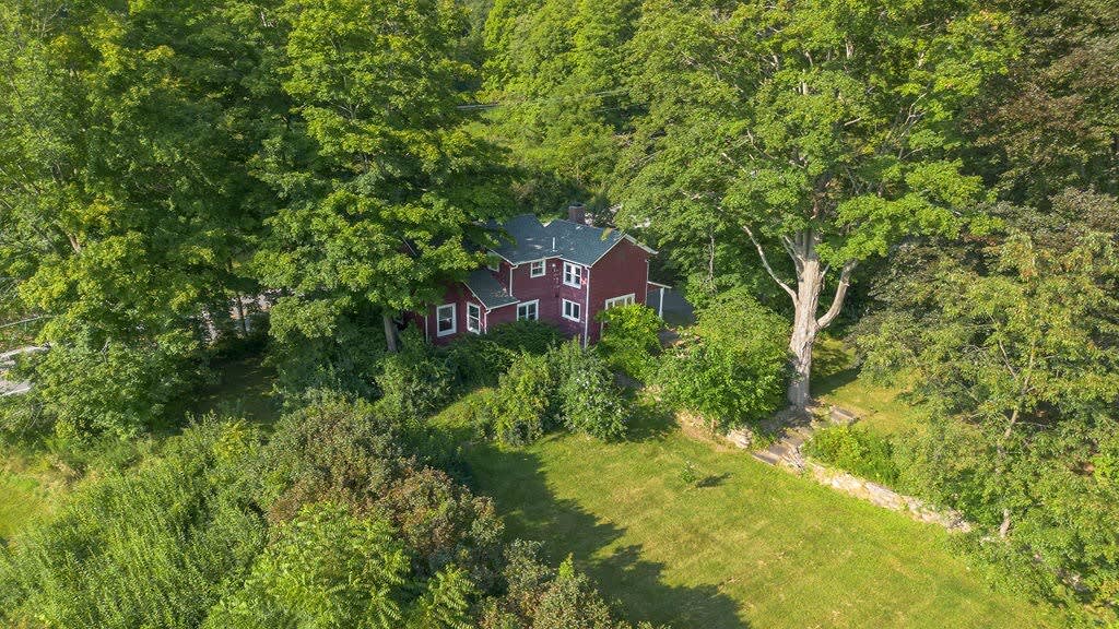 Aerial view of the red farmhouse nestled within a dense forest of mature green trees, showcasing its secluded rural setting. A large grassy area is visible to the right of the house.