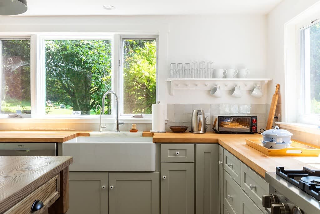 Kitchen sink area with a large white farmhouse sink, gooseneck faucet, and multiple windows overlooking lush greenery. Light gray cabinets, butcher block countertops, and open shelving for mugs complete the functional space.