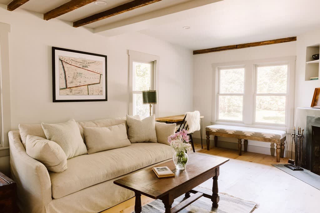 Inviting living room with a large, light-colored sofa adorned with throw pillows, a rustic wooden coffee table, and light wood floors. Exposed wooden beams and a framed map on the wall add character.