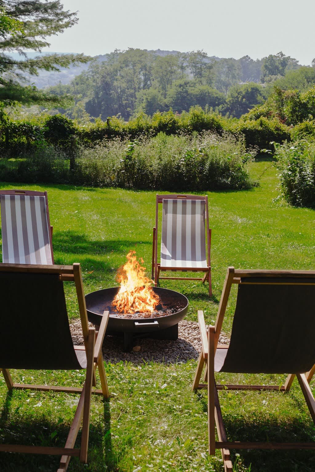 An outdoor fire pit with a roaring fire, surrounded by several lounge chairs on a gravel patch in a grassy yard. Lush green trees and a distant mountain view are visible in the background.