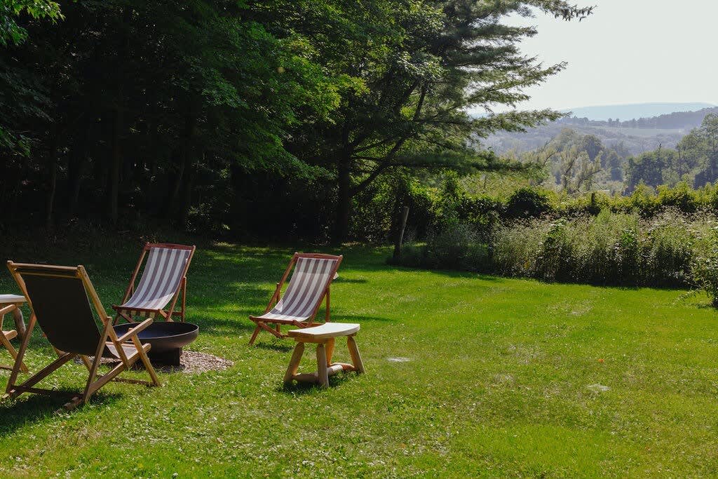 An expansive view of a well-maintained grassy yard with a fire pit and lounge chairs, offering a panoramic mountain view in the distance. Mature trees line the perimeter of the property.