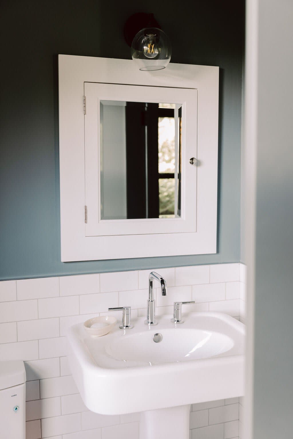 Bathroom vanity with a white pedestal sink, chrome faucet, and a white framed medicine cabinet mirror, set against a two-tone wall with white subway tiles on the bottom and a blue-gray paint above.