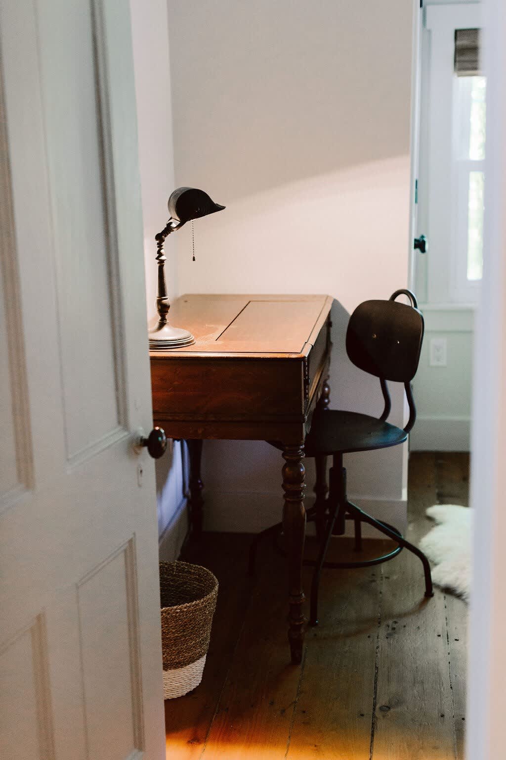 A vintage wooden desk with a black adjustable lamp and a matching black chair with a round back, set on hardwood floors. A woven basket sits on the floor next to the desk, and a white door is open to the left.