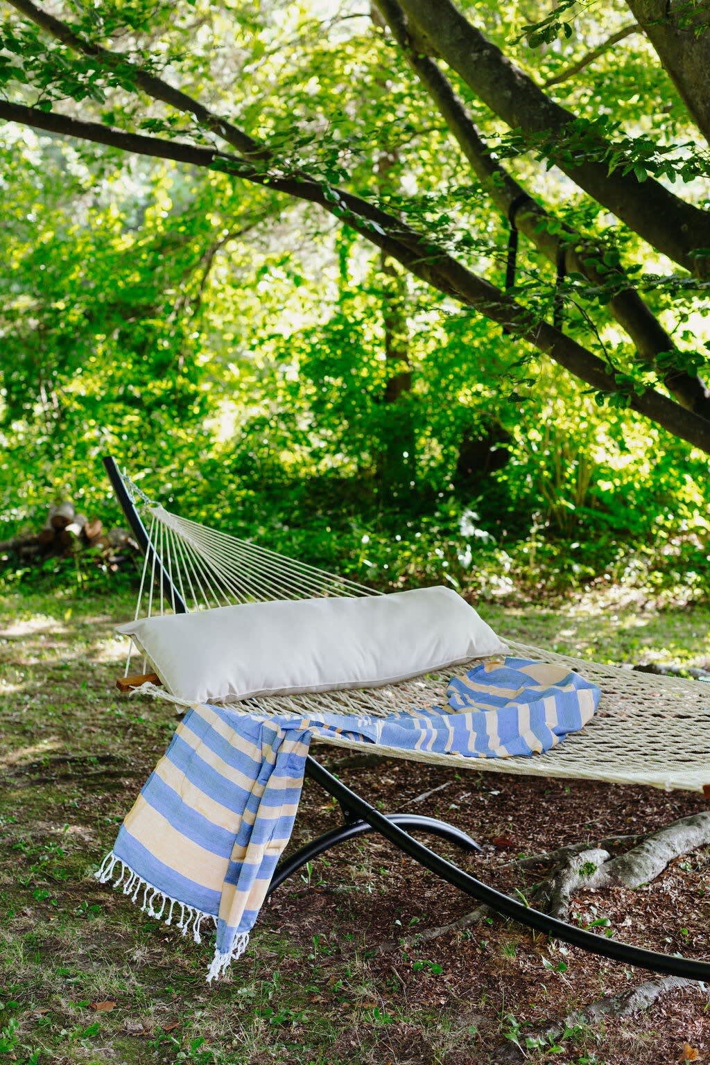 A comfortable rope hammock with a white pillow and a blue and yellow striped blanket is set up under the shade of large, leafy trees, offering a serene spot for relaxation in a natural, green environment.