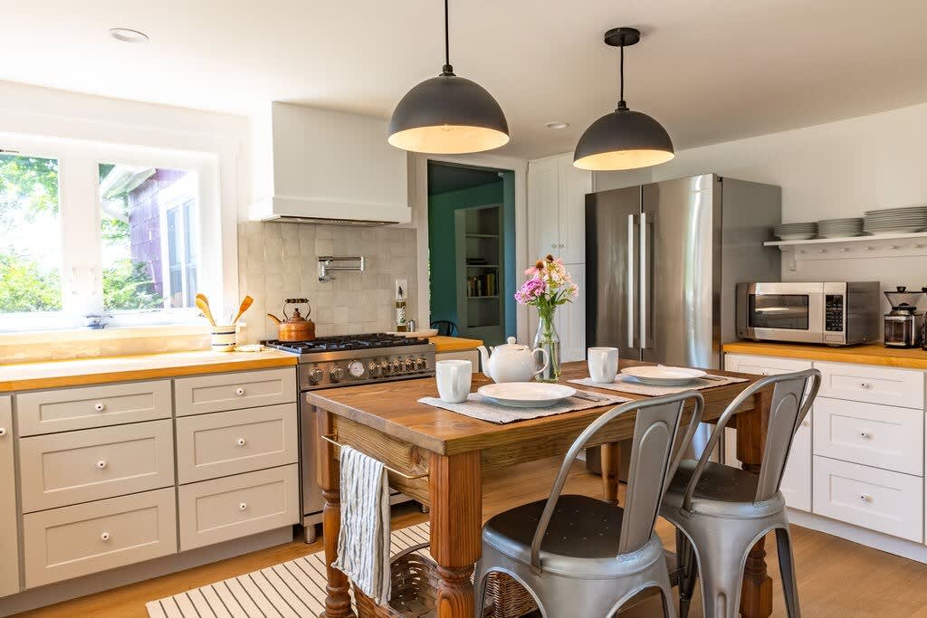 Spacious kitchen and dining area with a wooden island table set for two, industrial-style pendant lights, a stainless steel range, and a large refrigerator, all on light hardwood floors.