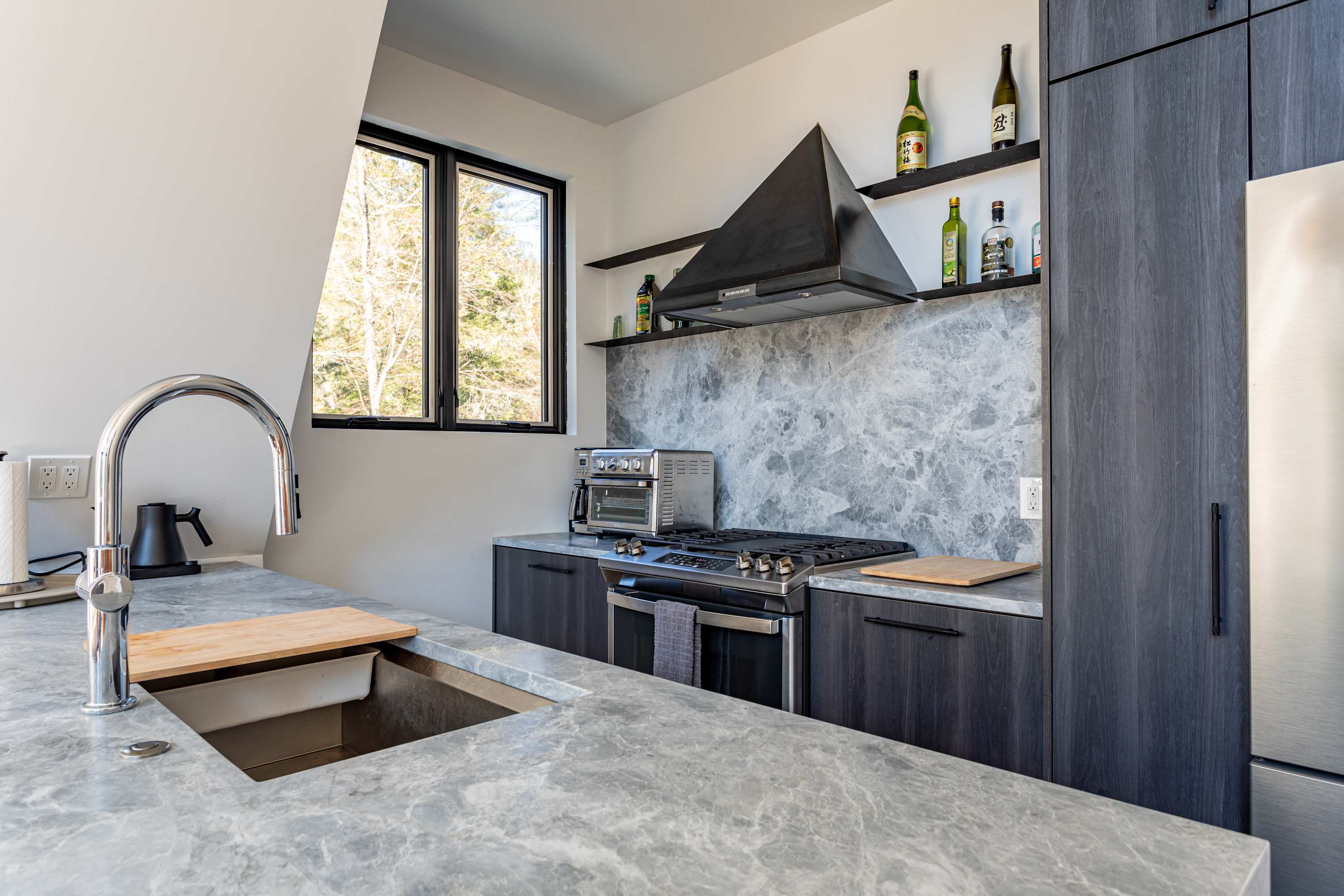 A close-up of the kitchen sink area, showcasing a modern stainless steel faucet, a large undermount sink, and gray marble-look countertops. A window above the sink offers a glimpse of the outdoors.