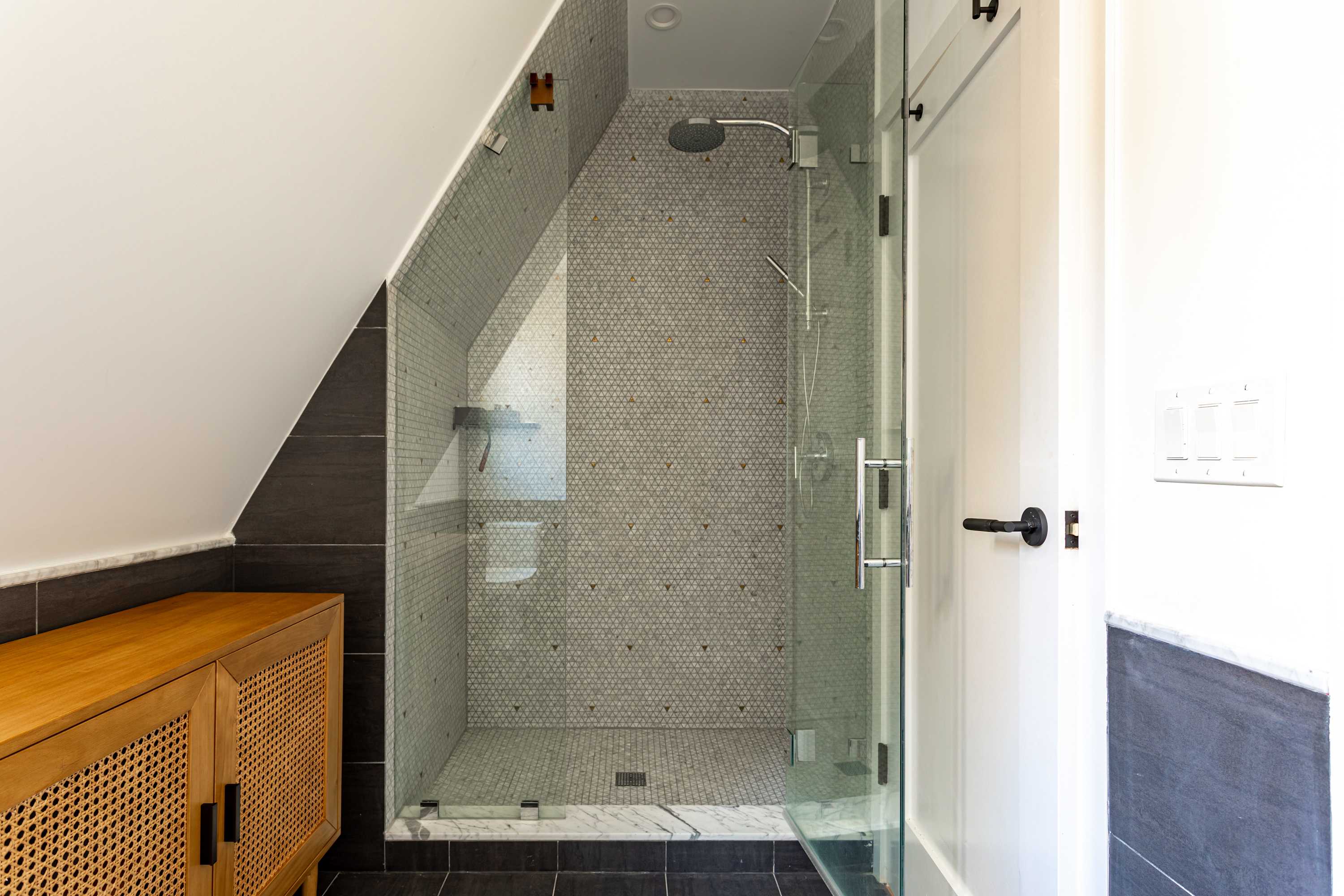 A spacious walk-in shower with glass enclosure and a rain showerhead, featuring geometric patterned tiles on the walls and a marble threshold. The shower is built into a slanted ceiling design.