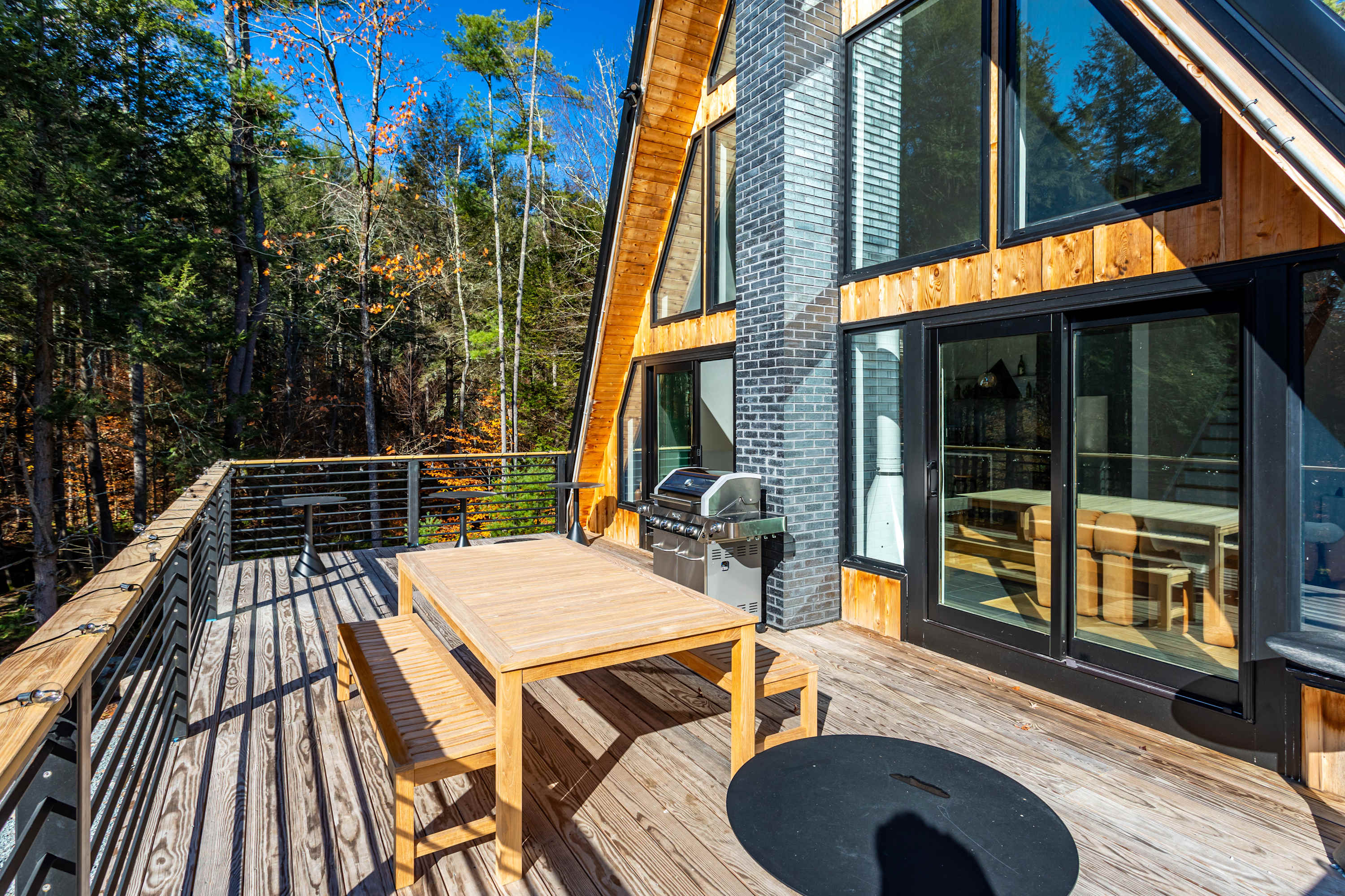A spacious wooden deck features an outdoor dining table with benches, a BBQ grill, and black metal railings, offering views of the surrounding forest through large glass doors of the A-frame house.