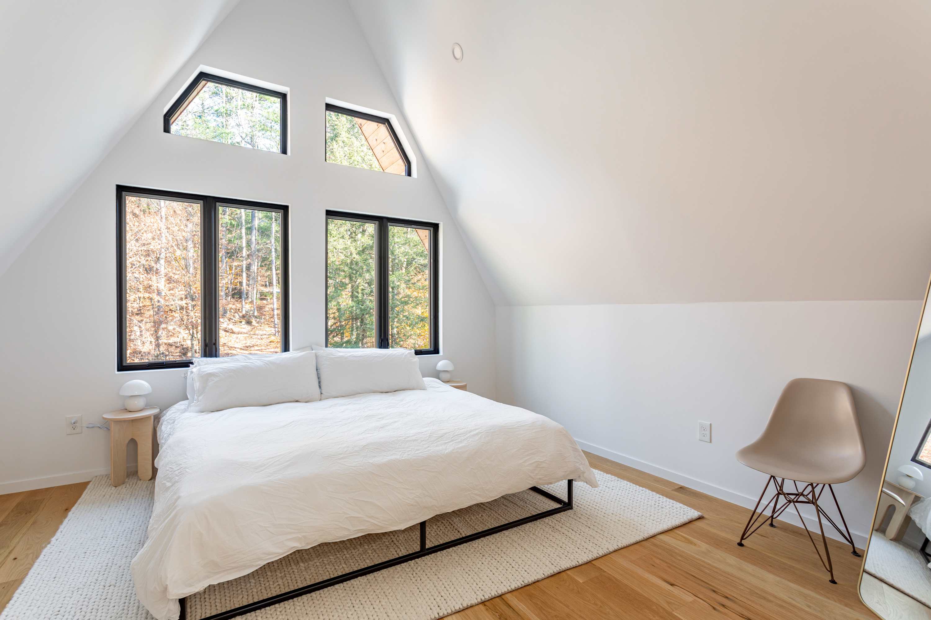 A bright bedroom with white walls and a high, sloped ceiling, featuring a queen bed with white bedding on a white shag rug. Multiple black-framed windows, including two triangular ones, offer views of the surrounding forest. A light-colored chair and small nightstands are visible.