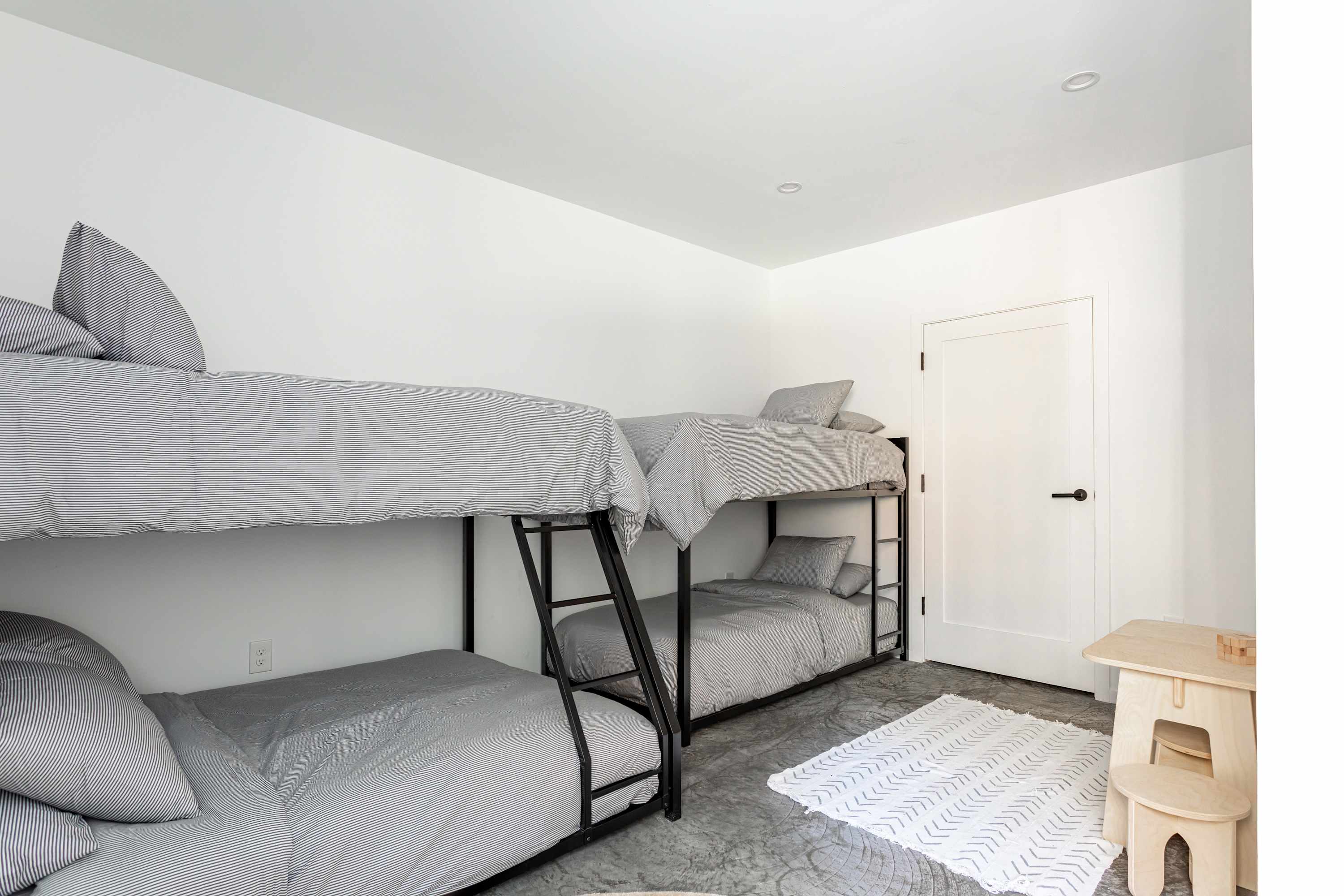 A minimalist bedroom with white walls features two black metal bunk beds with gray striped bedding, a small wooden table and chair set, and a white patterned rug on a concrete floor.