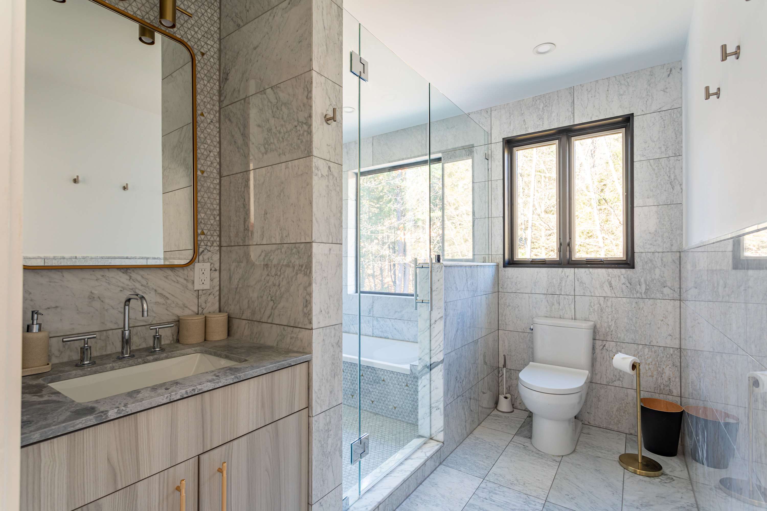 A modern bathroom with light gray marble-patterned tiles covering the walls and floor. It features a glass-enclosed shower, a white toilet, and a vanity with a light wood cabinet, marble countertop, and a gold-framed mirror. A window overlooks a wooded area.