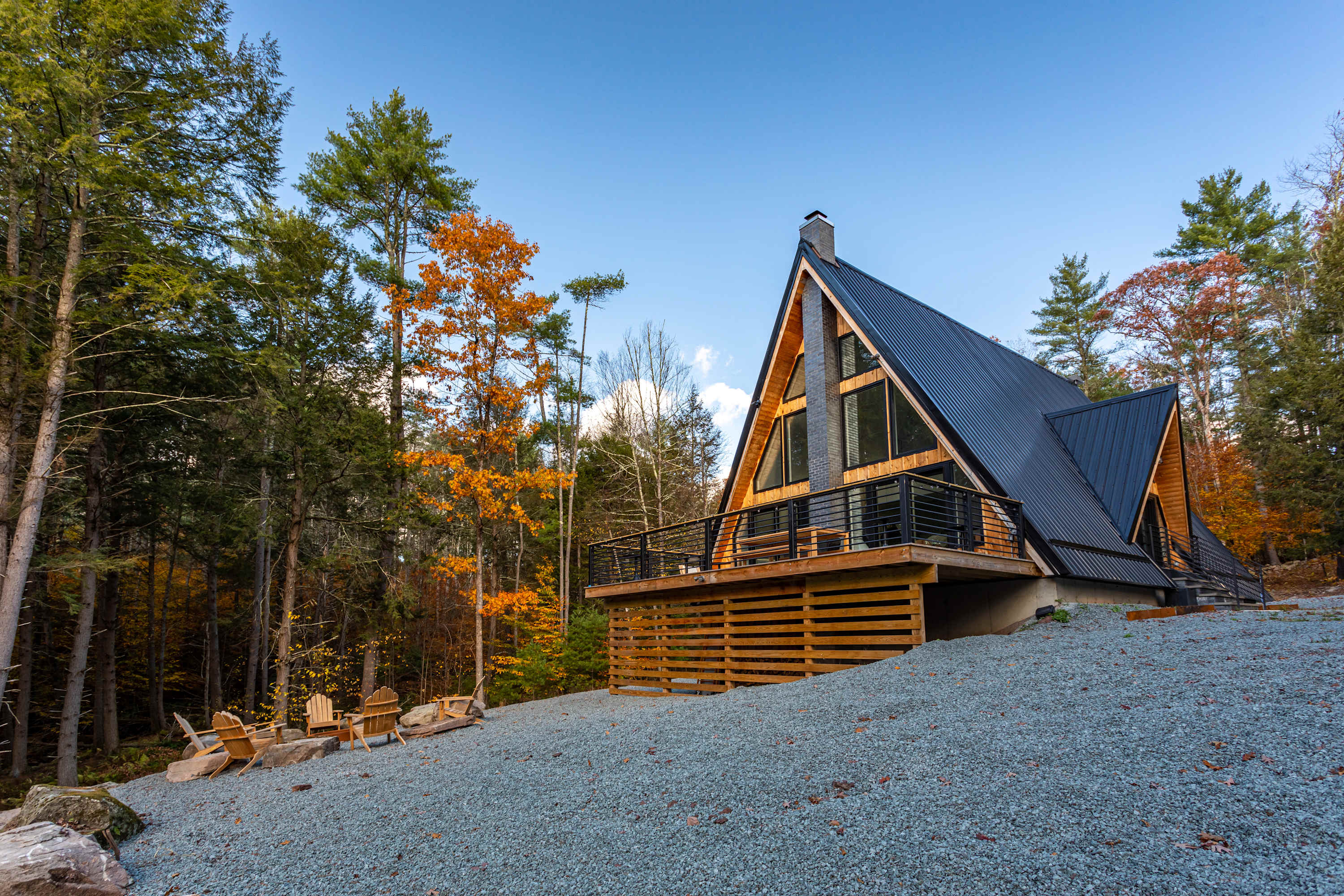 A modern A-frame house with a dark metal roof and natural wood siding is nestled among autumn trees, featuring a large wooden deck with black railings and outdoor seating on a gravel landscape.