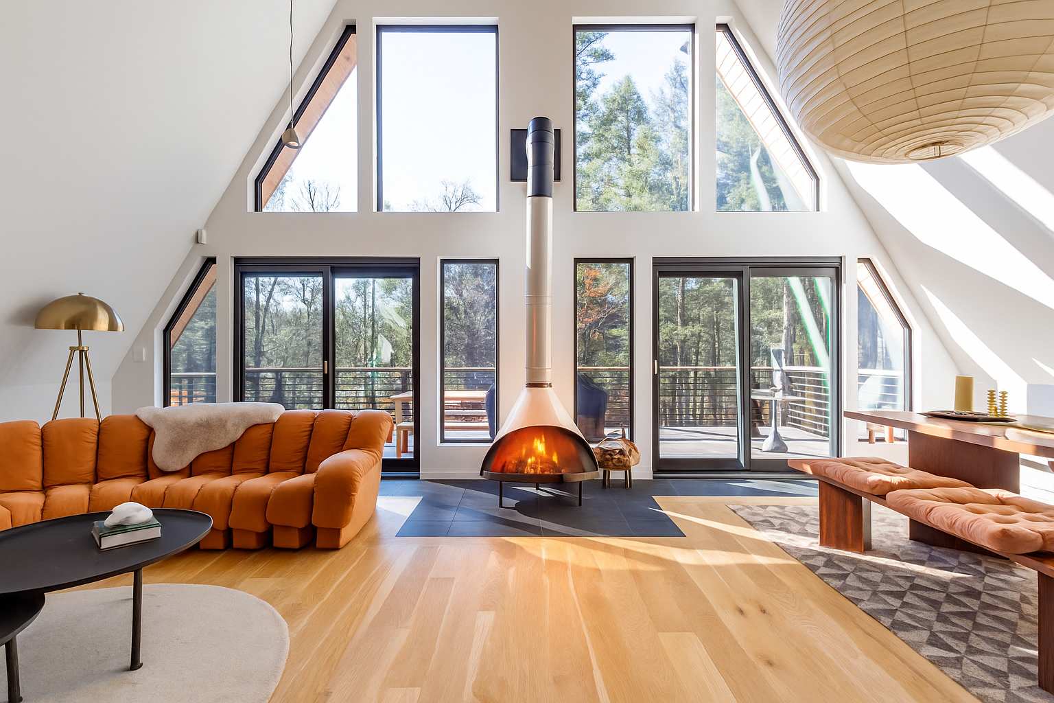 Expansive living room with a large orange modular sofa, a modern freestanding fireplace, and floor-to-ceiling A-frame windows offering forest views. Hardwood floors extend to a dining area.