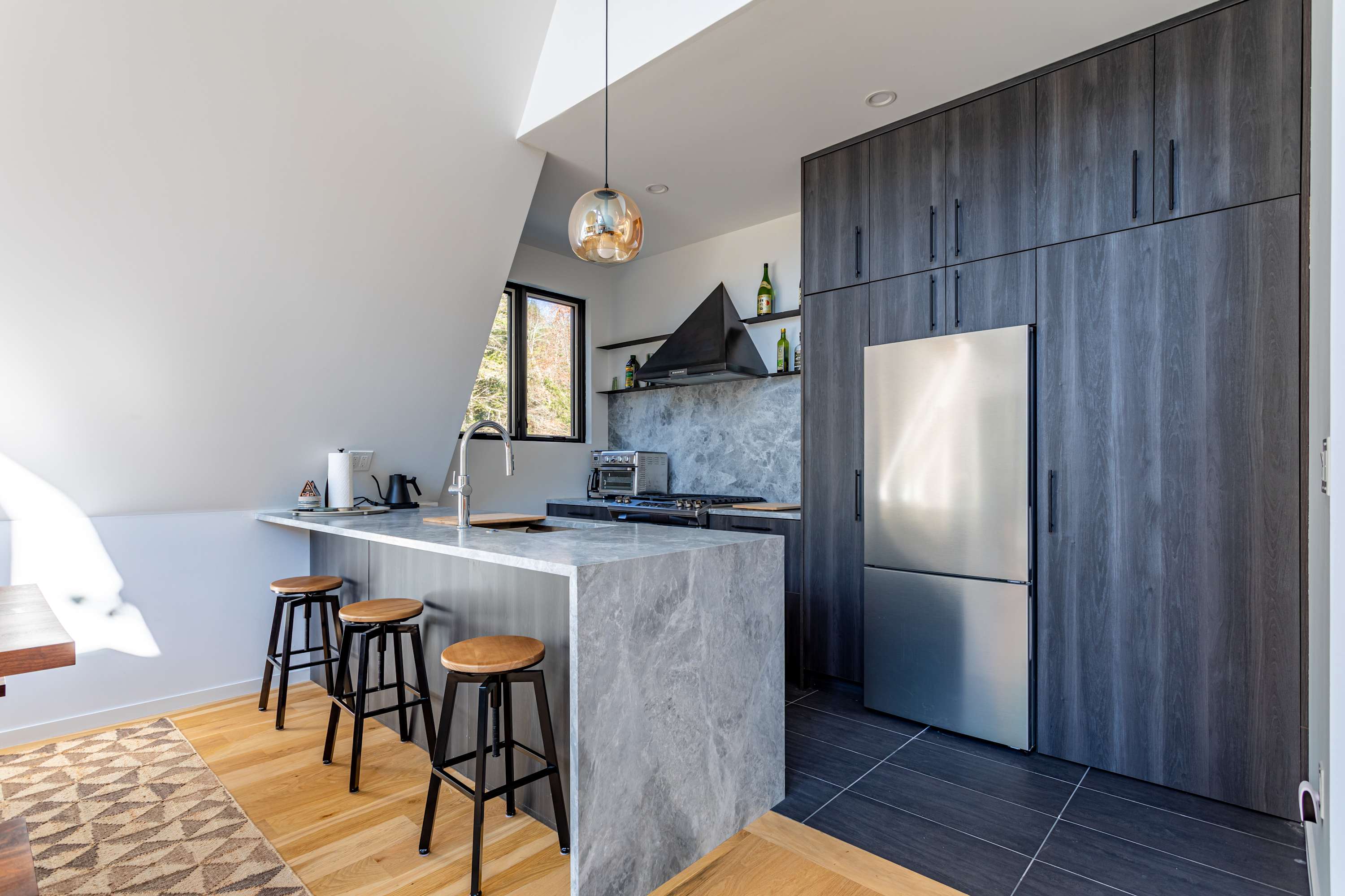 A modern kitchen with dark wood-grain cabinetry, a gray marble-look countertop and backsplash, and stainless steel appliances. A kitchen island with bar stools provides additional seating.