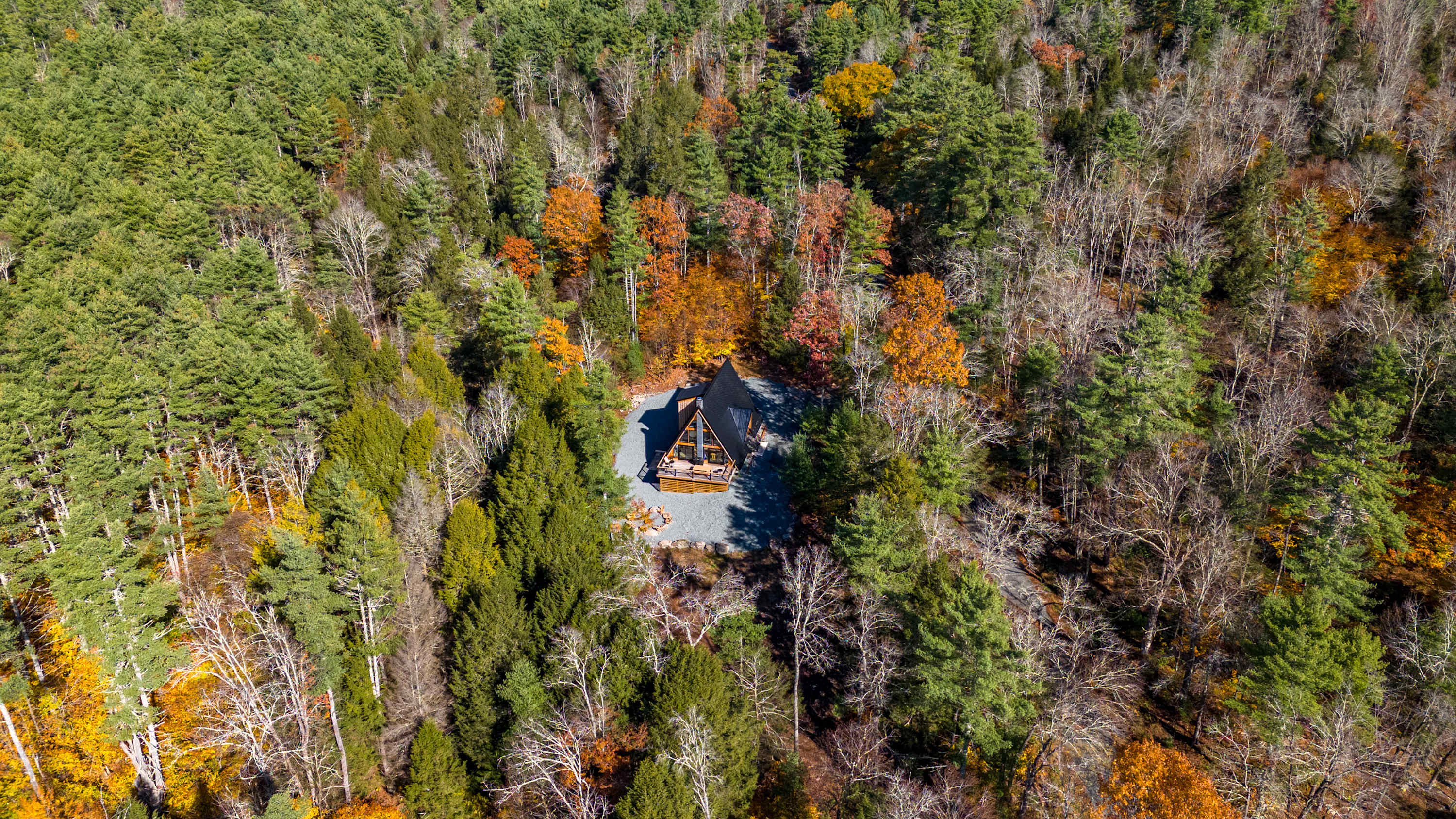 Aerial view of the A-frame house nestled in a dense forest, surrounded by a mix of evergreen and deciduous trees displaying autumn colors. A gravel driveway leads to the property.