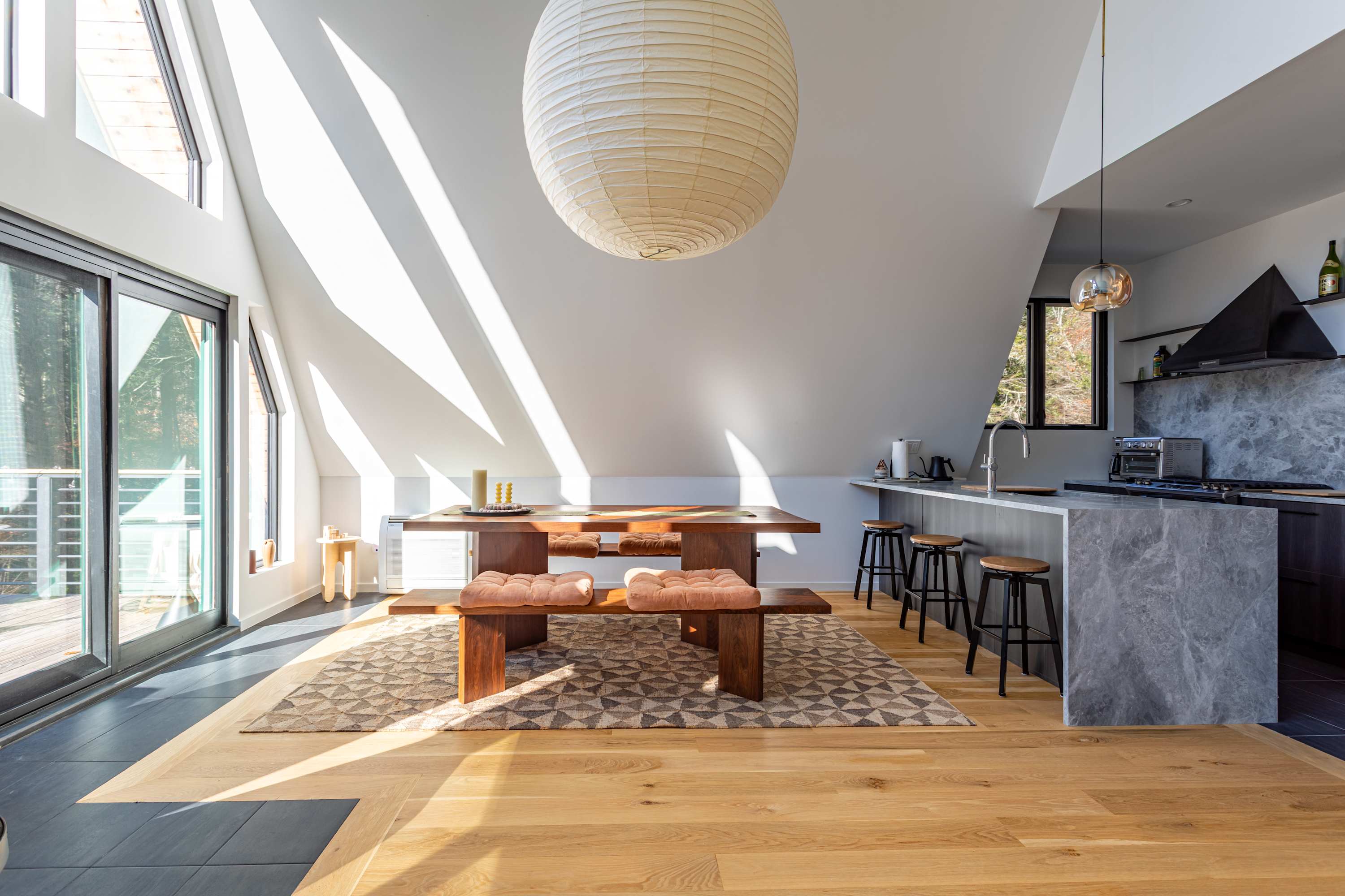 Sunlit dining area with a large wooden table and matching benches, set on a geometric rug. Adjacent to a modern kitchen with grey marble counters and dark cabinetry, all under a large paper lantern.