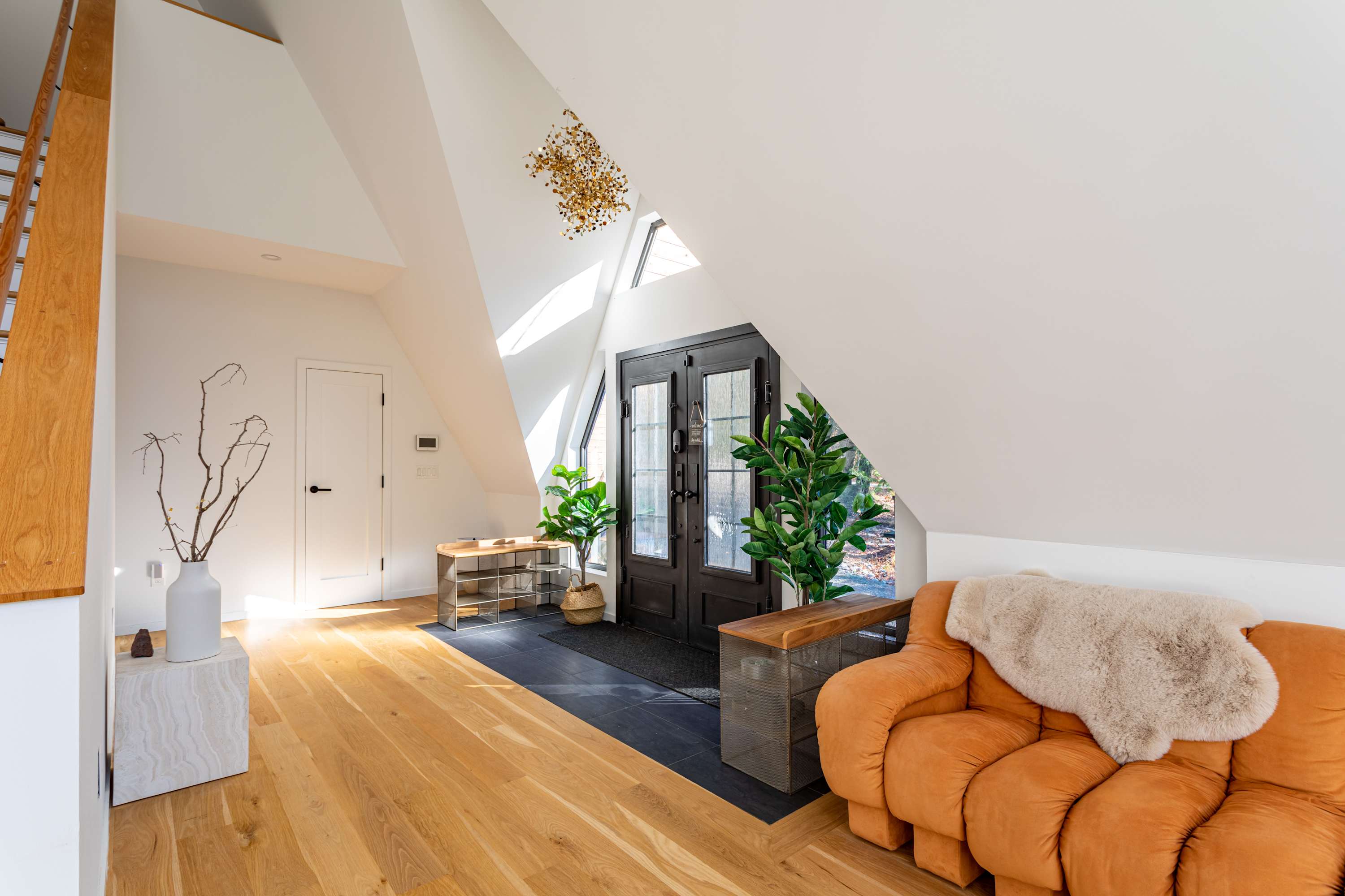 Bright foyer with double black doors, hardwood floors, and a modern orange sofa. The high, angled ceilings and large windows create an airy, welcoming entrance.