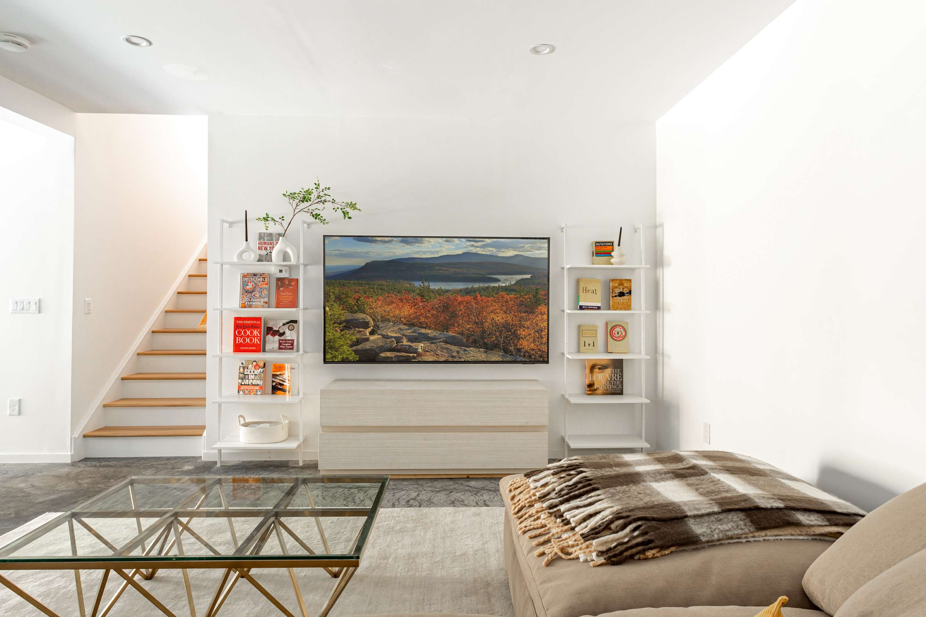 A bright living room with polished concrete floors and white walls, featuring a large TV centered between white ladder-style bookshelves. A neutral L-shaped sectional sofa with a plaid throw blanket and a glass coffee table are arranged on a light rug. A staircase is visible to the left.