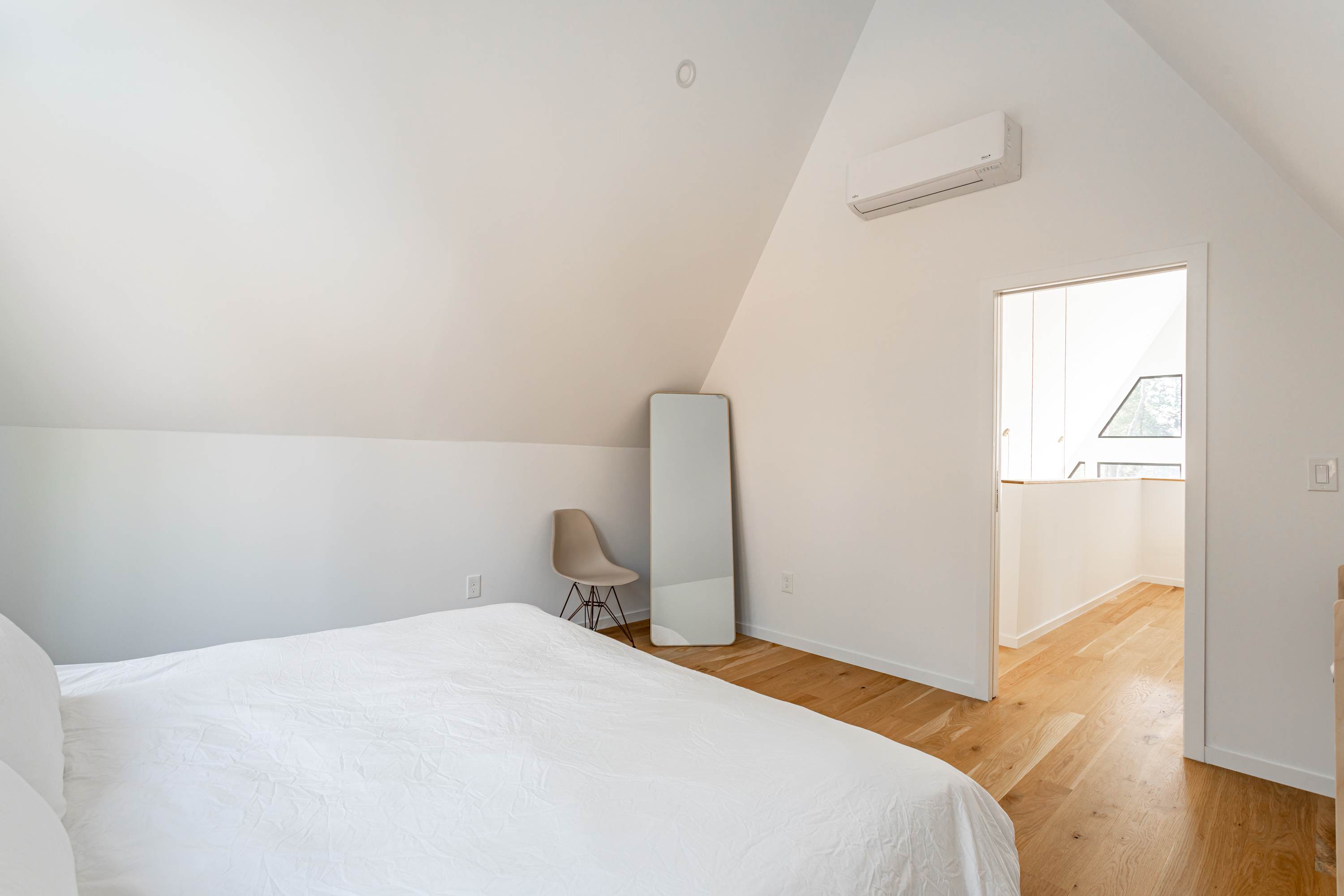 A minimalist bedroom with white walls and a sloped ceiling, featuring a queen bed with white bedding. A light-colored chair and a full-length mirror lean against the wall. An air conditioning unit is mounted high on the wall, and a doorway leads to another area.