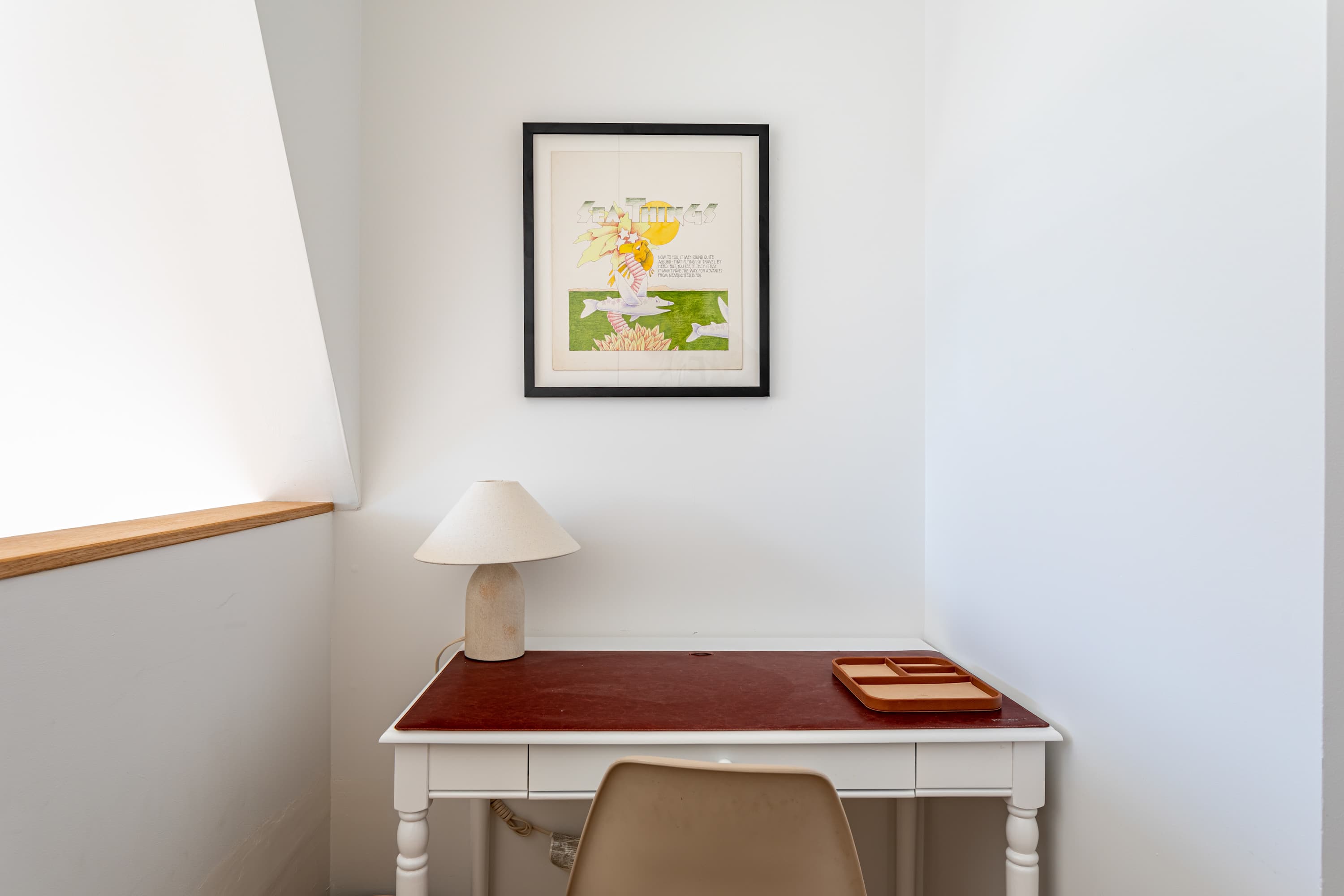 A cozy corner workspace features a white desk with a red leather top, a cream-colored lamp, and a framed art print on the white wall, illuminated by natural light from an angled window.