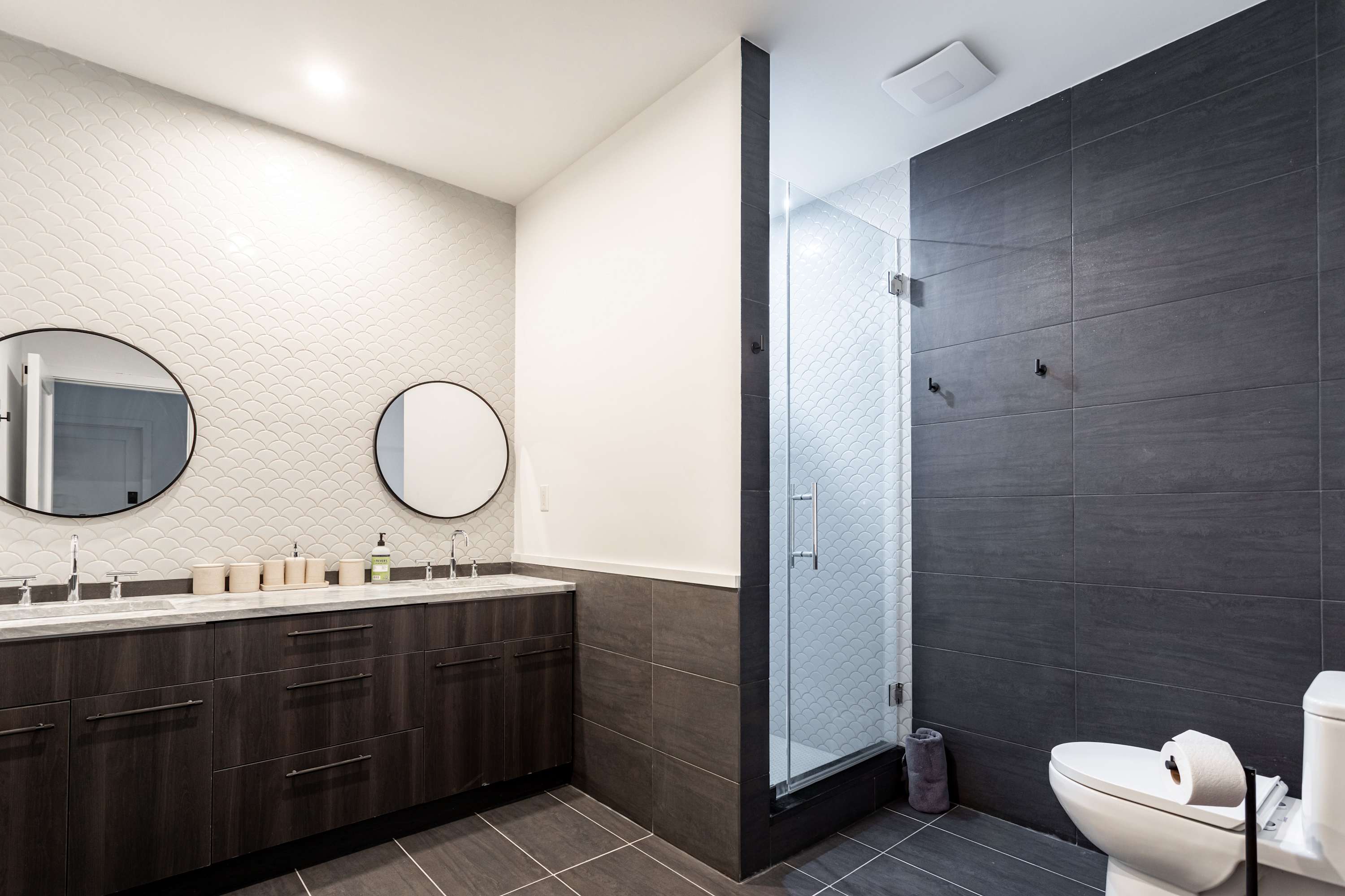 A modern bathroom features a dark wood double vanity with two round mirrors, a white scalloped tile accent wall, and a spacious walk-in shower with dark gray large format tiles and a glass enclosure.