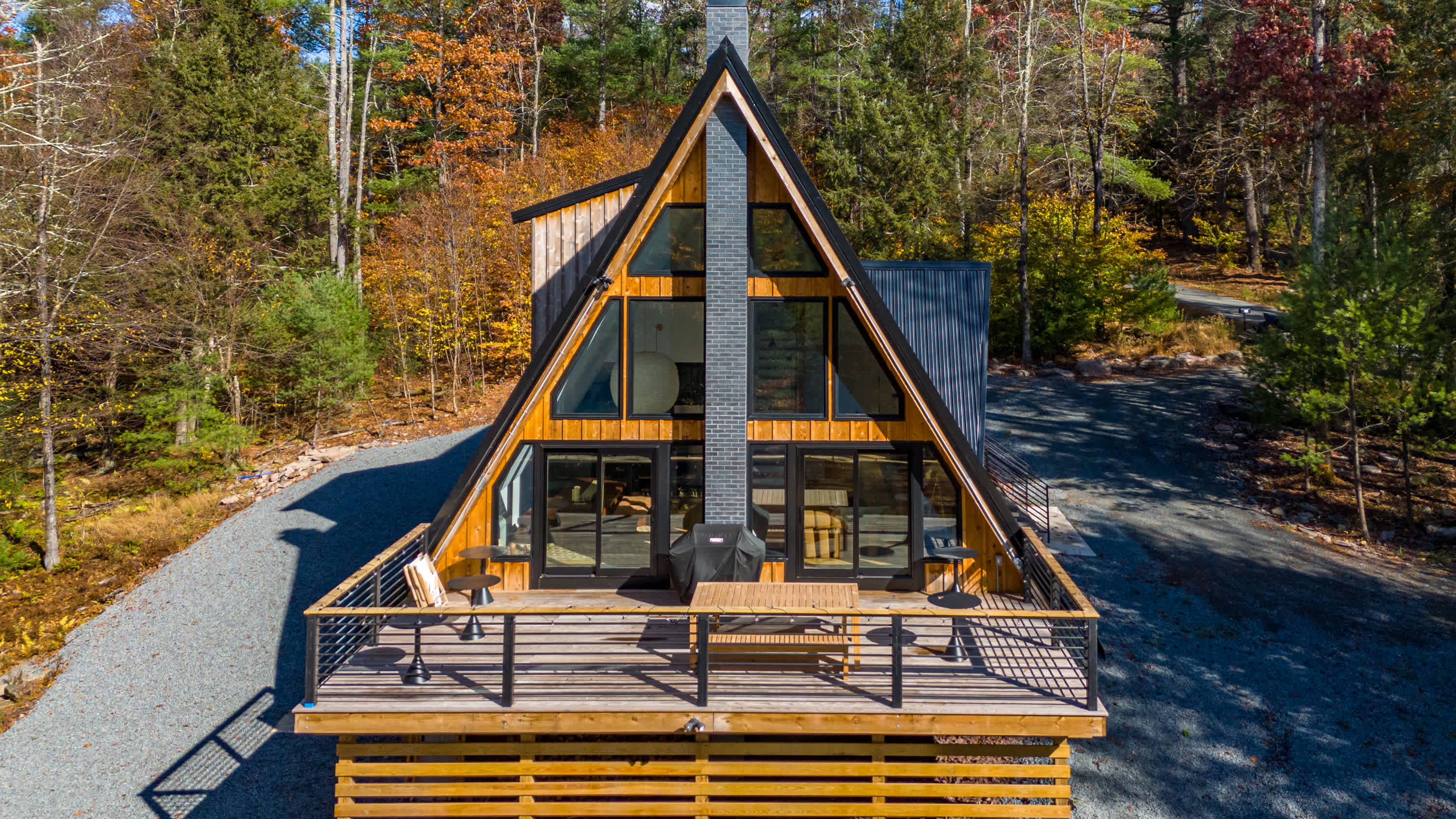An aerial view showcases the striking A-frame architecture of the house with its tall chimney and large windows, a spacious wooden deck with a grill and outdoor dining, surrounded by vibrant autumn foliage.