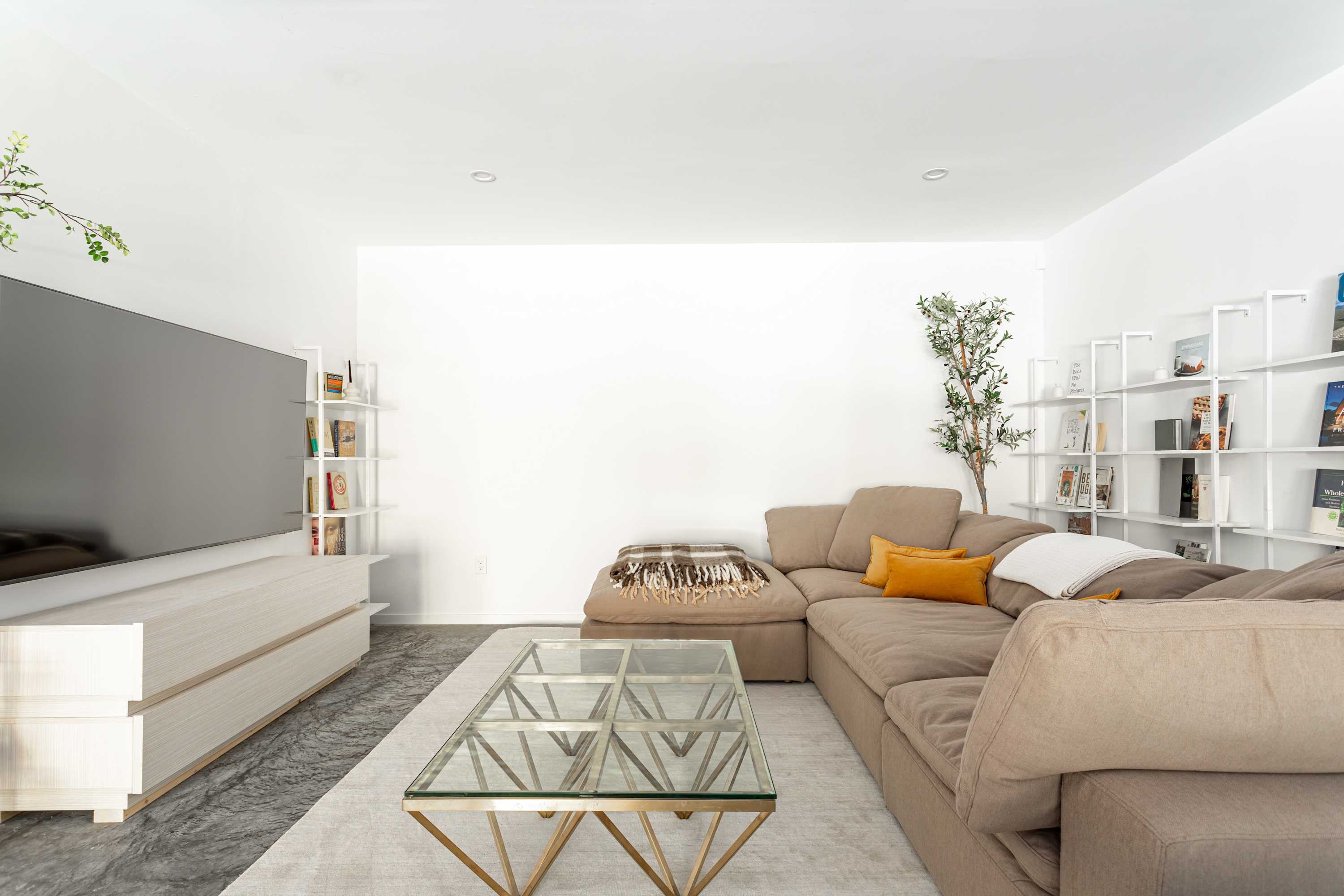 A spacious living room with dark gray polished concrete floors and white walls. A large L-shaped sectional sofa in a neutral tone is adorned with throw pillows and a blanket. A glass coffee table sits on a light rug, and a large TV is mounted above a low console, flanked by white shelving units.